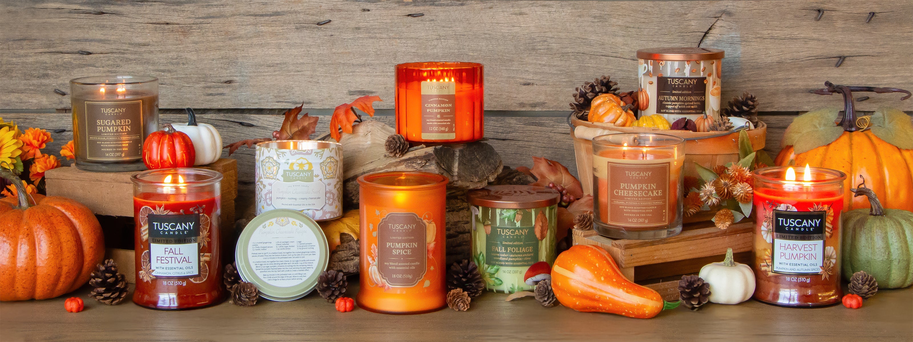 Three Tuscany Candle jars—Cinnamon Spiced Café, Fall Festival, and Pumpkin Cheesecake—displayed with pumpkins and autumn leaves in front of a window with a fall scene.