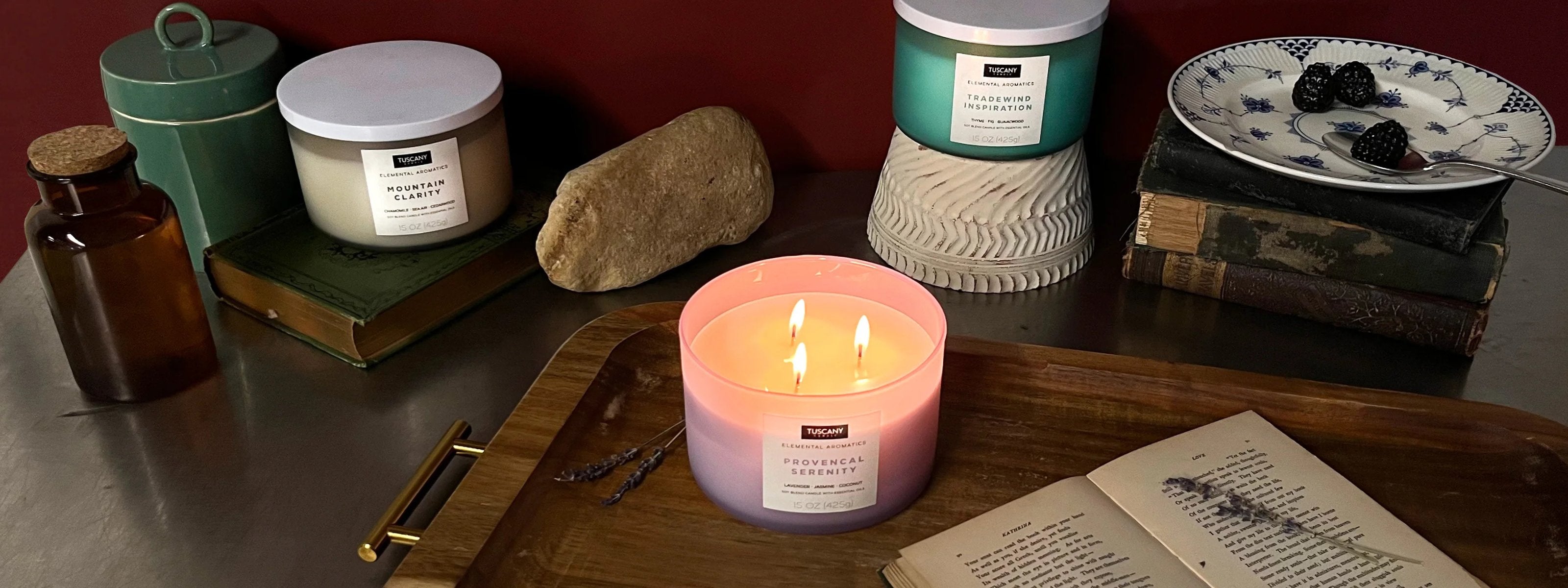 A lit candle on a tray with a book, candle snuffer, and dried flowers. Nearby are two closed candles, a rock, a cup, and blackberries on books against a red wall.