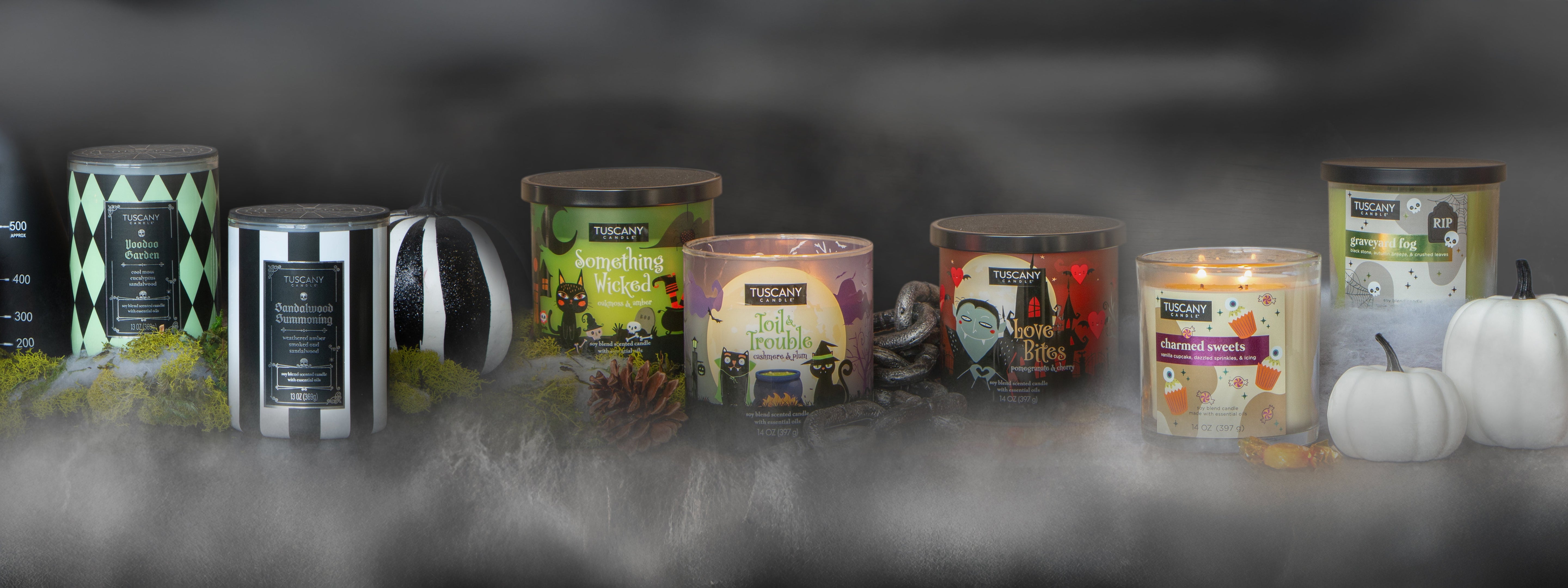 A row of Tuscany Candle brand Halloween-themed candles and jars, surrounded by decorative pumpkins, pine cones, and moss, on a foggy surface.