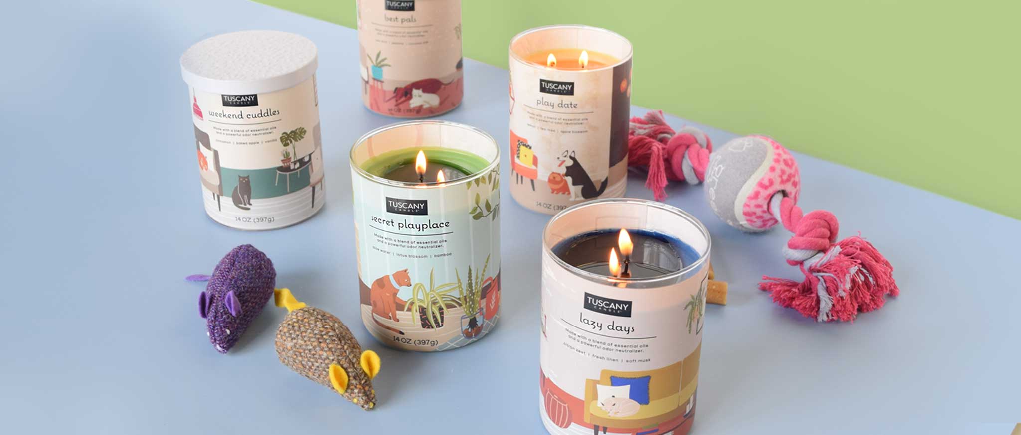 Four scented candles in glass jars are lit and surrounded by pet toys, including two plush mice and a rope toy, on a light blue and green surface.