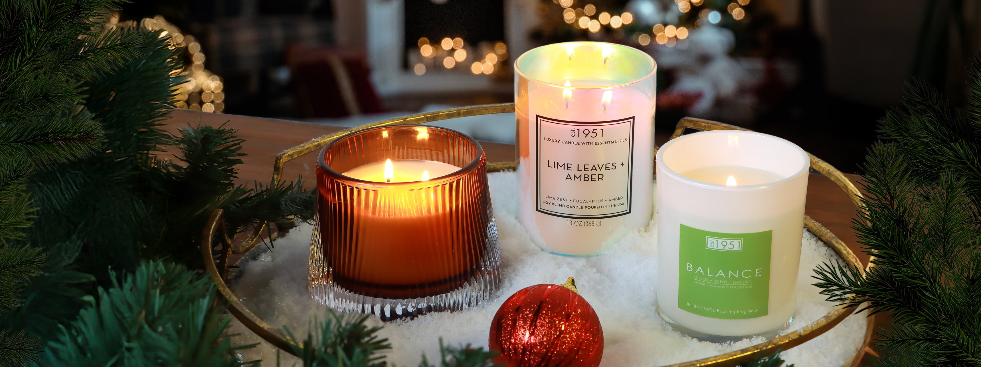Three lit candles in glass holders sit on a tray with artificial snow, green pine branches, and a red ornament, with festive lights blurred in the background.