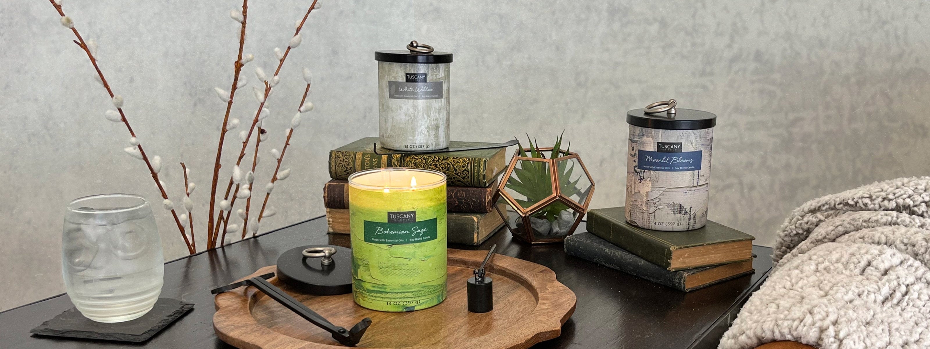 Candles on a table surrounded by stacked books, a glass tumbler, a small plant, and a brown textured throw blanket.