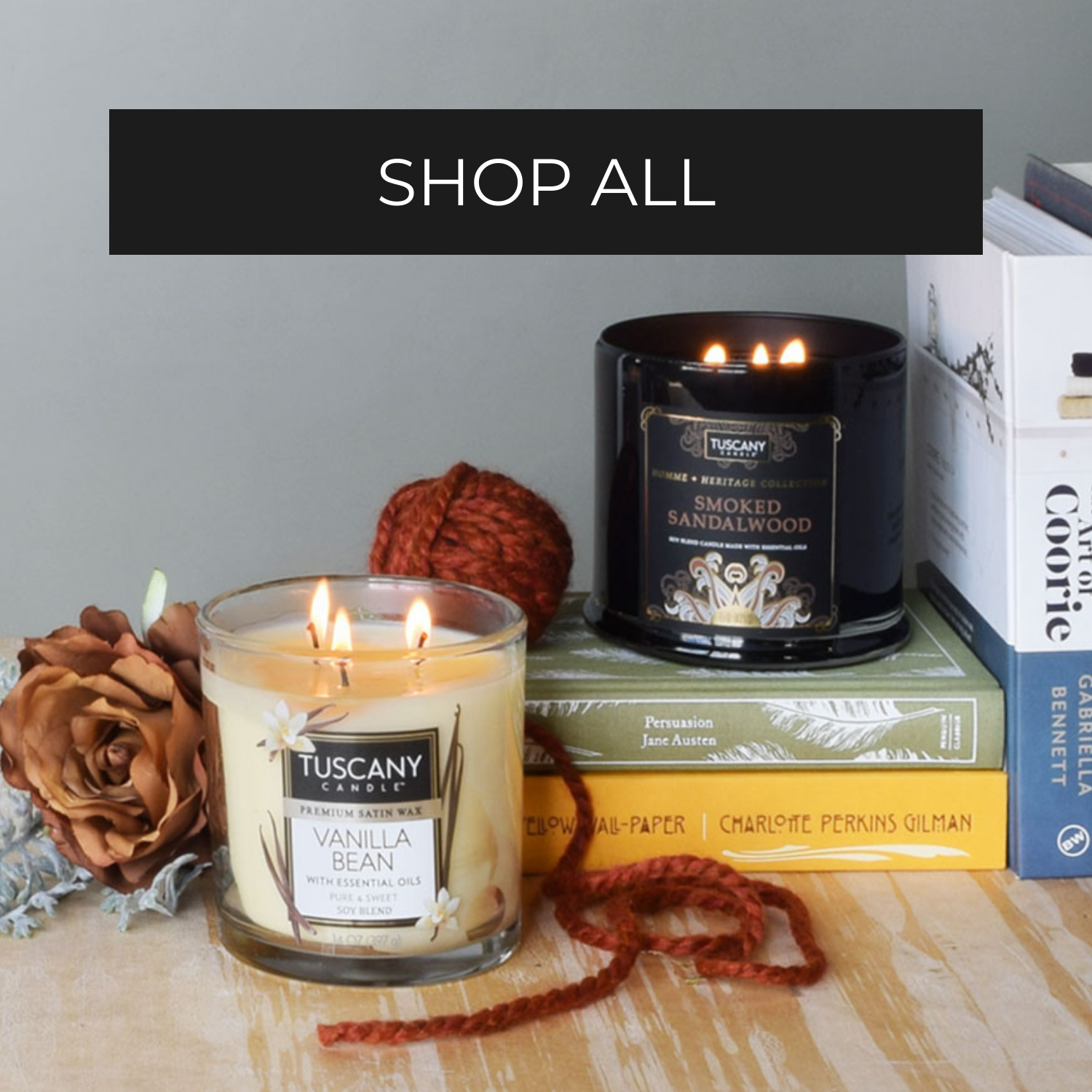 Two Tuscany Candle jars, one Vanilla Bean and one Smoked Sandalwood, are displayed on stacked books with a brown flower, yarn, and the text "SHOP ALL" above.
