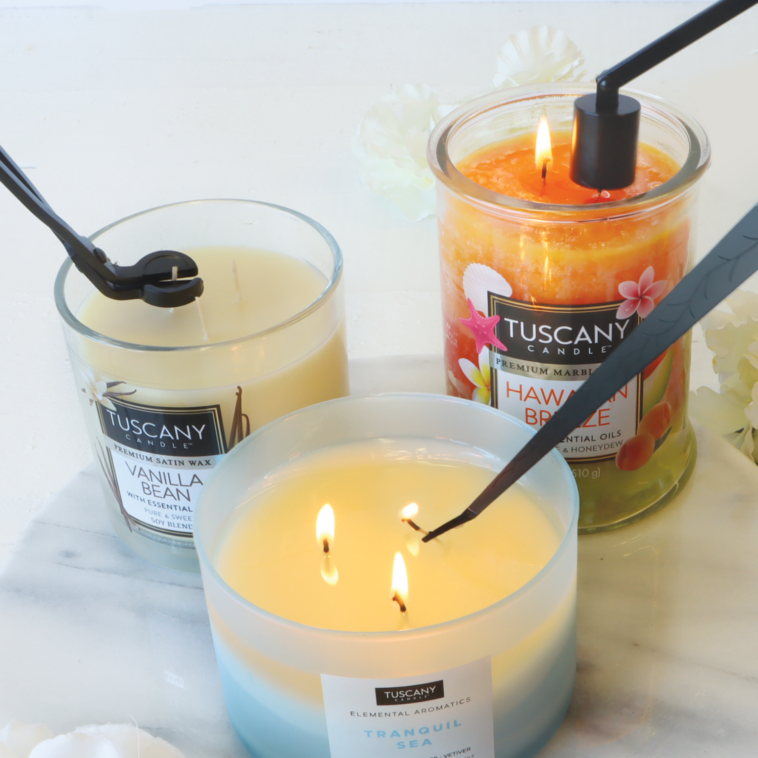 Three lit candles on marble, gently extinguished by a snuffer from the Tuscany Candle® SEASONAL 3-Piece Candle Care Set, release delightful scents: Vanilla Bean, Hawaiian Breeze, and Tranquil Sea—a scene perfect for those who cherish maintaining an aromatic ambiance.
