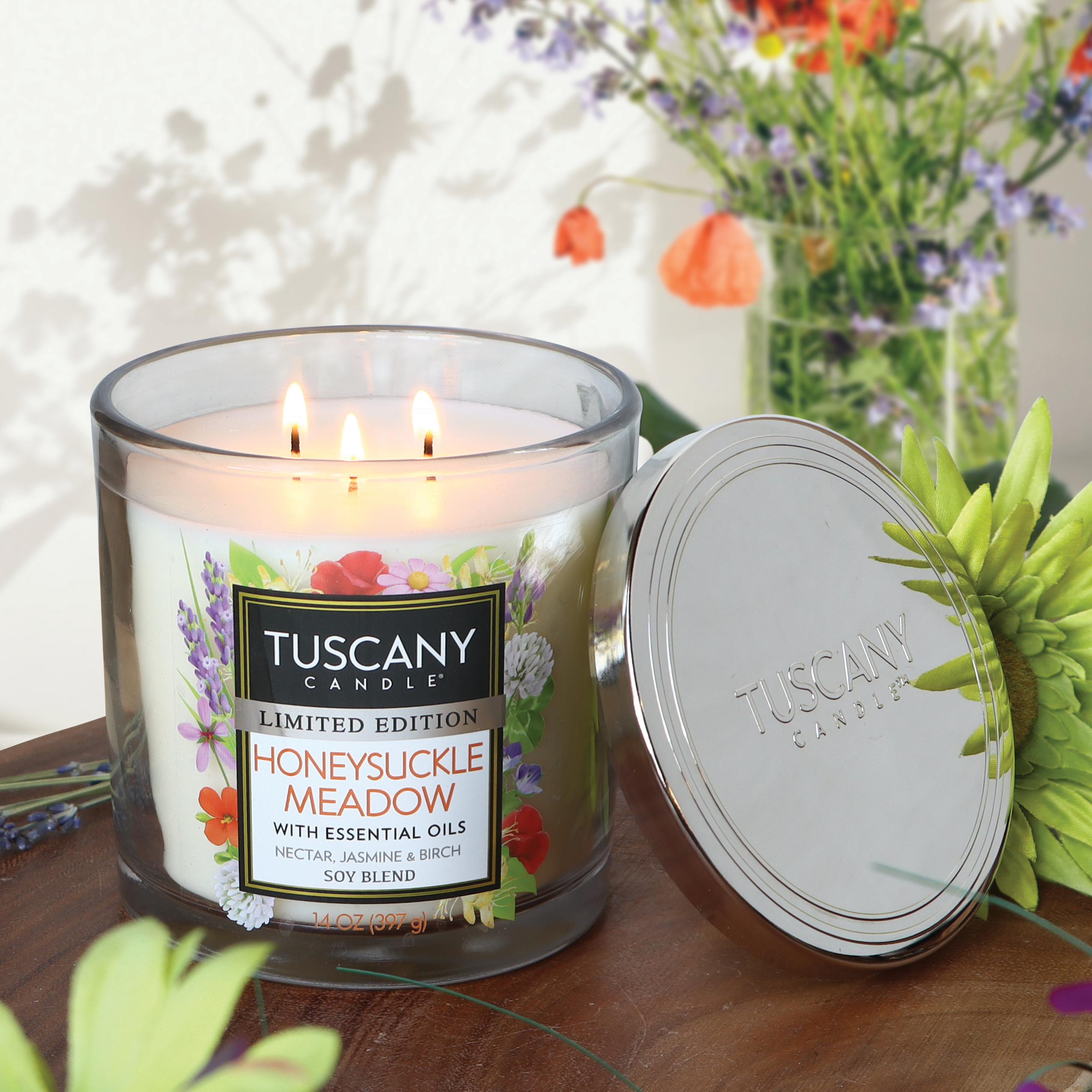 A Honeysuckle Meadow scented candle on a table next to a bouquet of soft florals. (Tuscany Candle® SEASONAL)