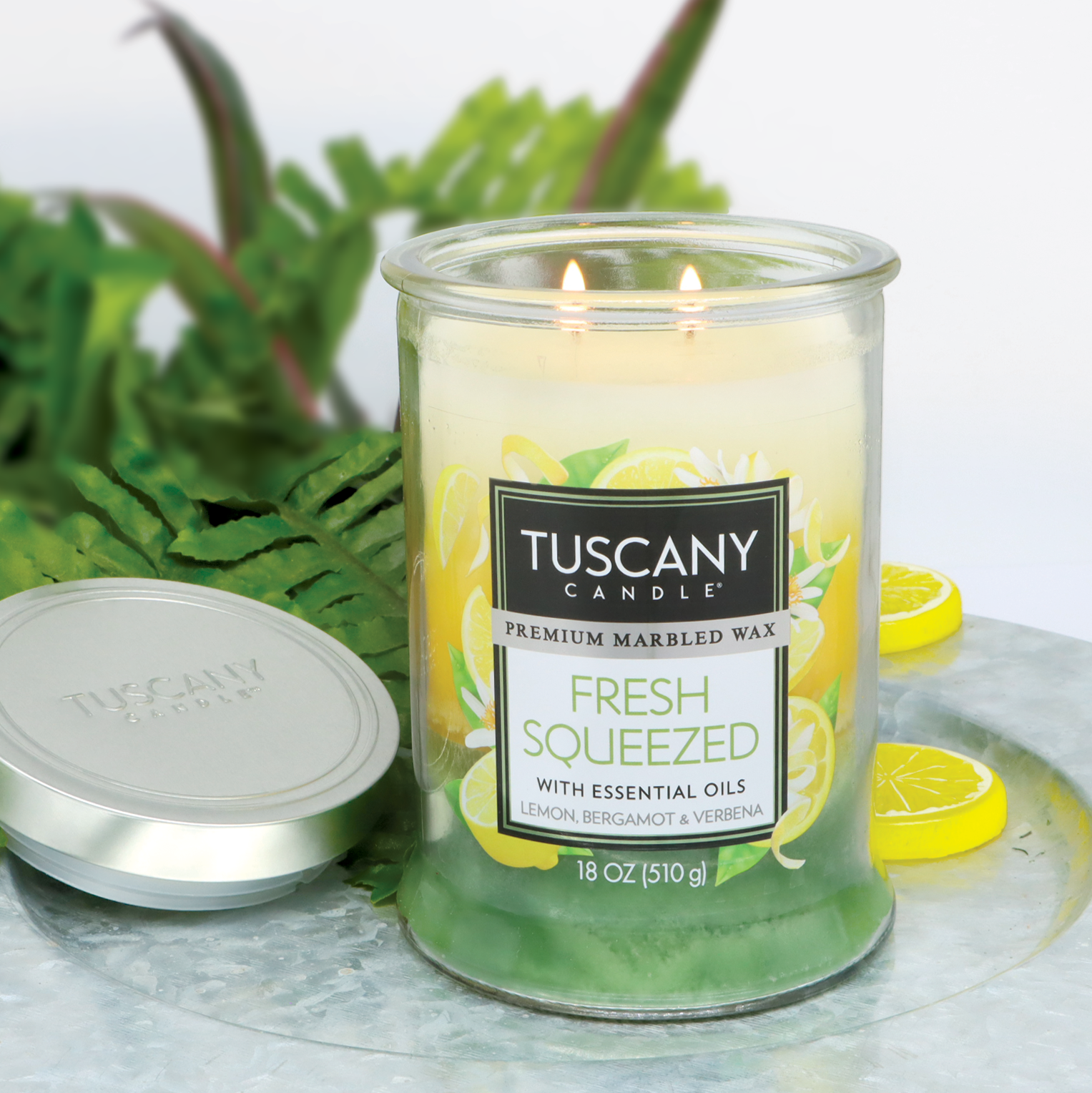 An 18 oz Tuscany Candle® EVD Fresh Squeezed Long-Lasting Scented Jar Candle with a lemon bergamot verbena design sits on a table, lit with two wicks. Green leaves decorate the background, and its lid is nearby.