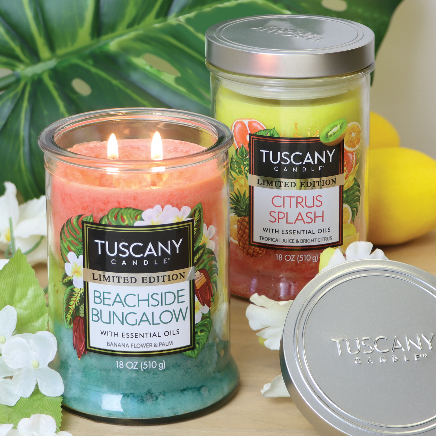 A Tuscany Candle® SEASONAL "Beachside Bungalow: Banana Scented Summer Candle" (18 oz) and a "Citrus Splash" candle are on a table with white flowers and green leaves, with one candle lit.