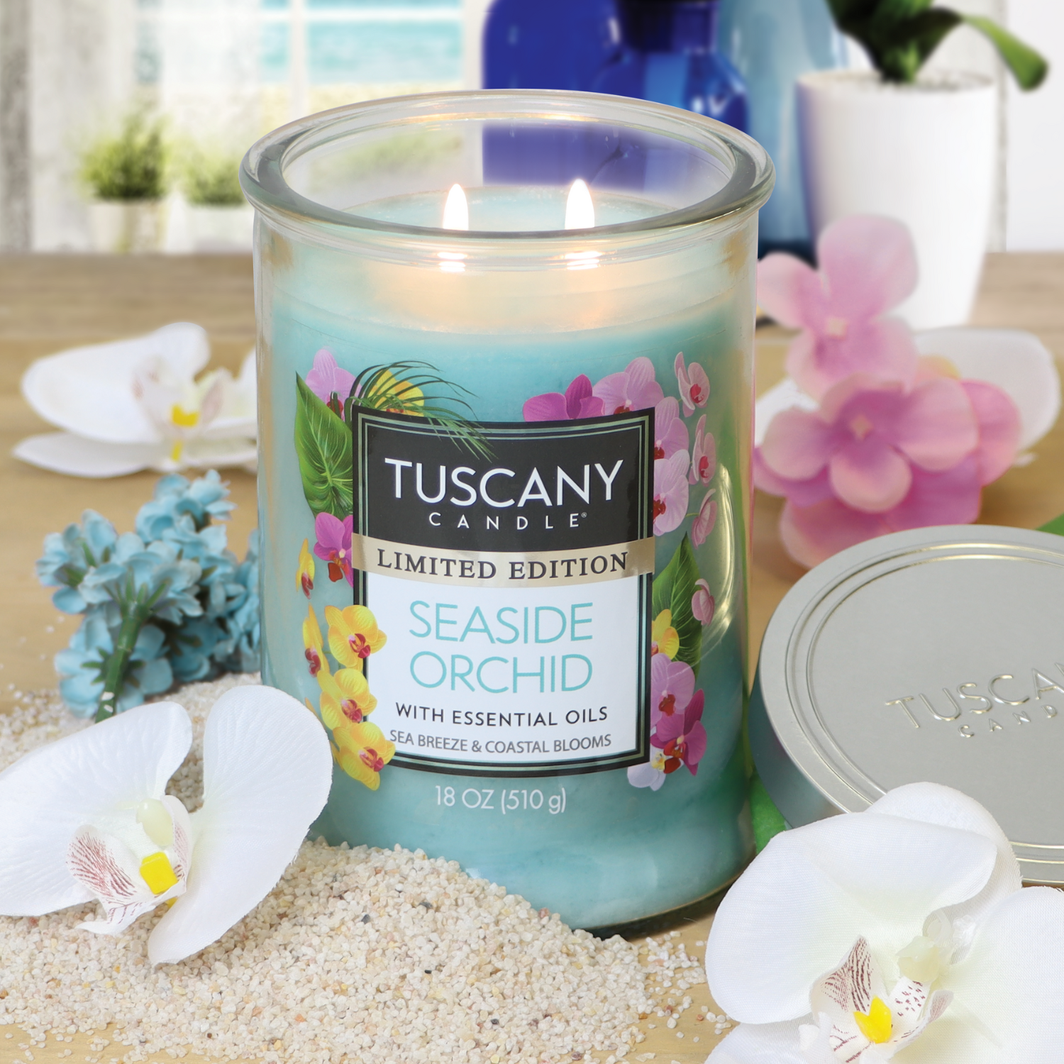 A lit Tuscany Candle® SEASONAL "Seaside Orchid: Coastal Scented Summer Candle (18 oz)" rests on sand with seashells, white and pink flowers, and a decorative blue bottle in the background.