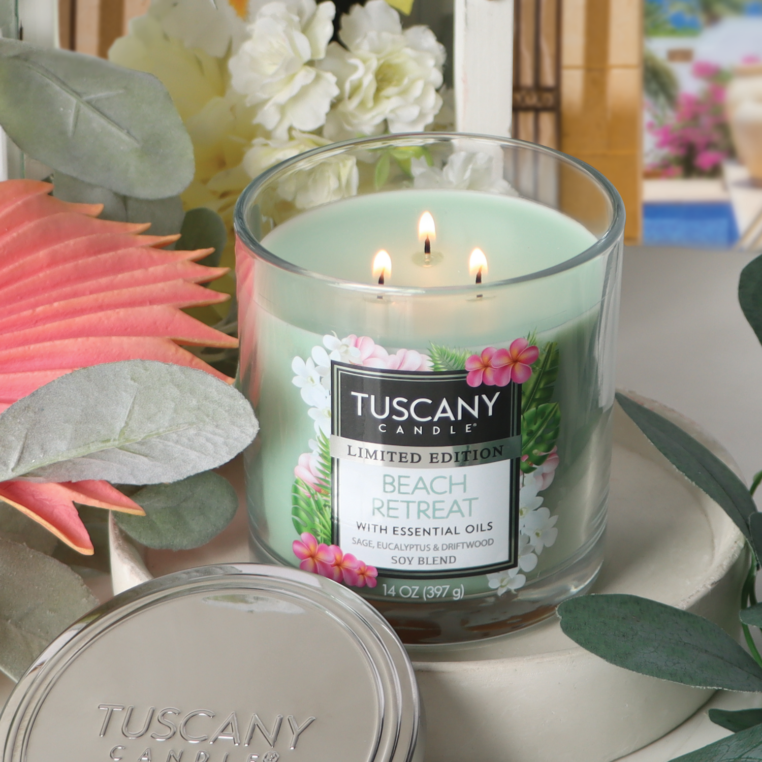 A lit candle labeled "Beach Retreat: Eucalyptus & Sage Scented Summer Candle" (14 oz) by Tuscany Candle® SEASONAL, in a glass jar surrounded by leaves and flowers.