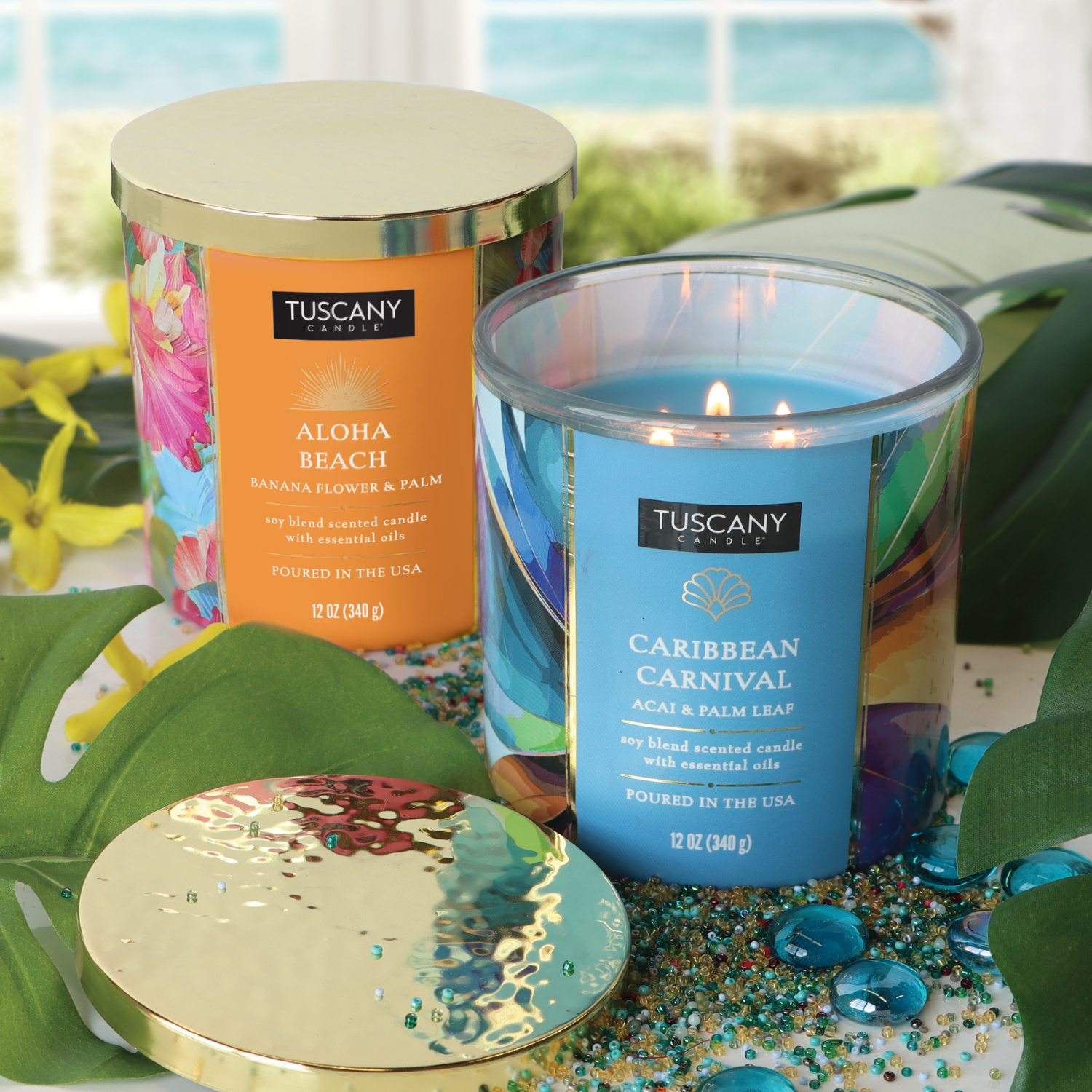 Tuscany Candle® SEASONAL’s Aloha Beach: Banana Scented Summer Candle (12 oz) from the Island Glam Collection sits among lush leaves and beads, with a beach view—creating the perfect tropical summer candle display.