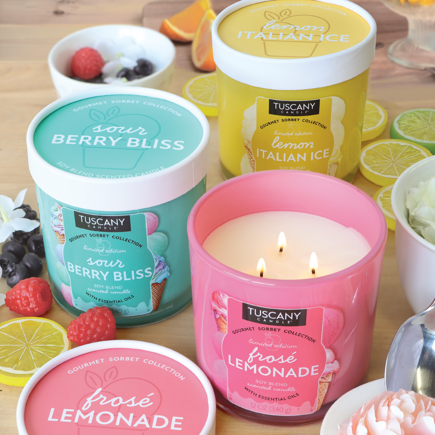 Three 12 oz Tuscany Candle® SEASONAL Gourmet Sorbet Collection candles, labeled Sour Berry Bliss (Blue Raspberry Scent), Lemon Italian Ice, and Frosé Lemonade, are displayed on a table with various fruits.