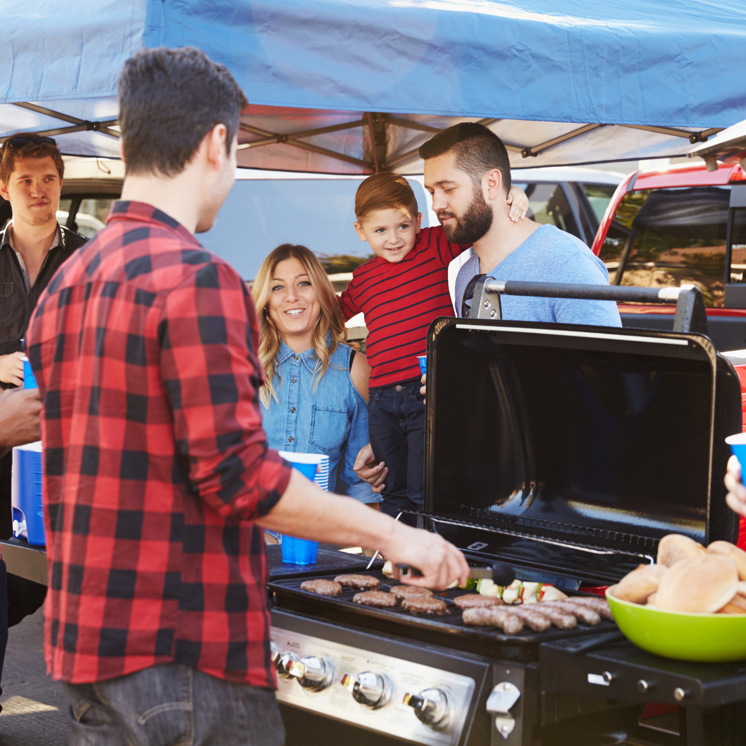 At an outdoor gathering under a canopy, people grill food and enjoy the warm aroma of the Tailgate Scented Football KC Heart Candle (13 oz) by Tuscany Candle x Charlie Hustle, while an adult holds a child nearby.