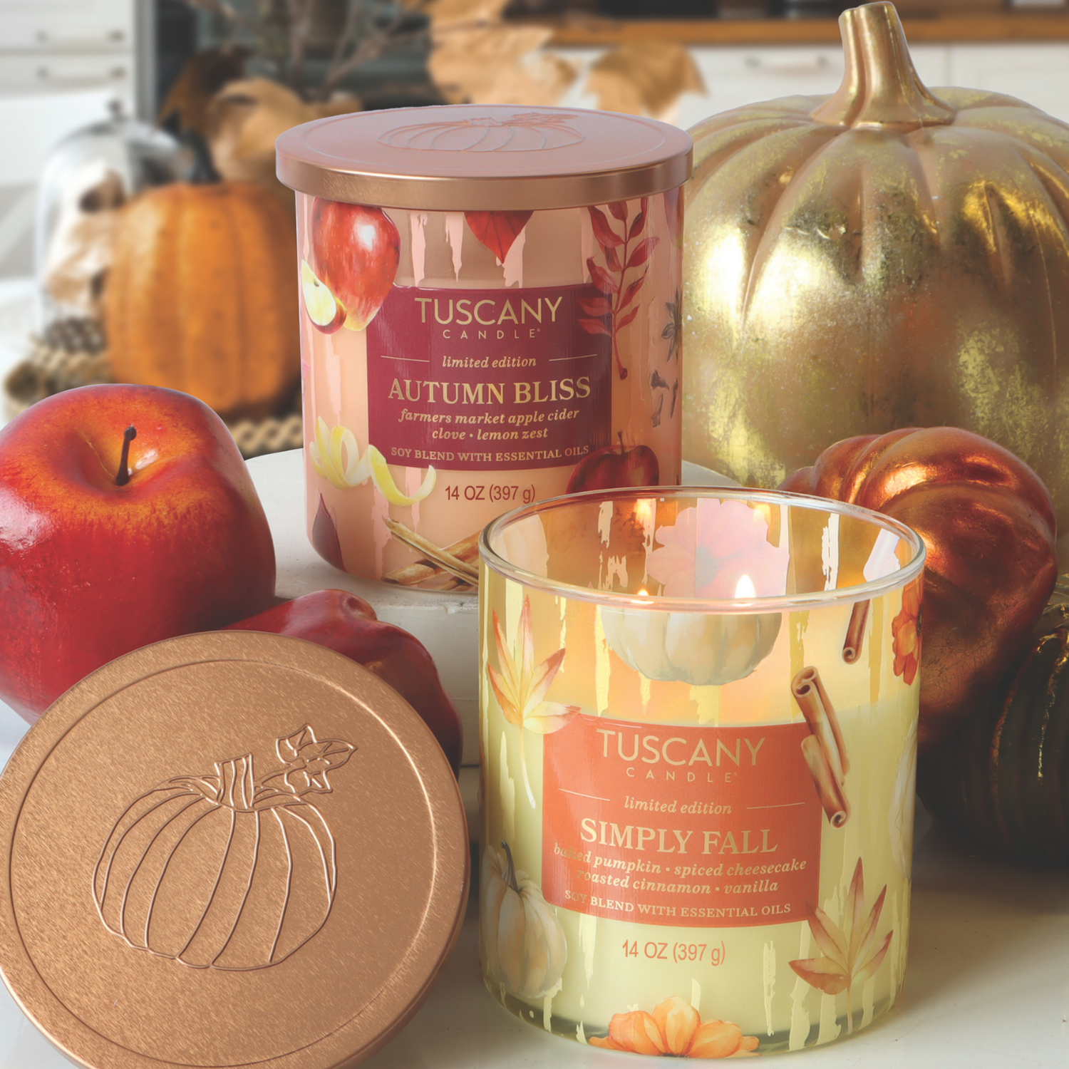 Two Tuscany Candle® SEASONAL jars—Autumn Bliss: Apple Cider & Clove Scented Fall Candle (14 oz) from the Simply Fall Collection—are on a table with artificial apples, pumpkins, and fall décor in the background.