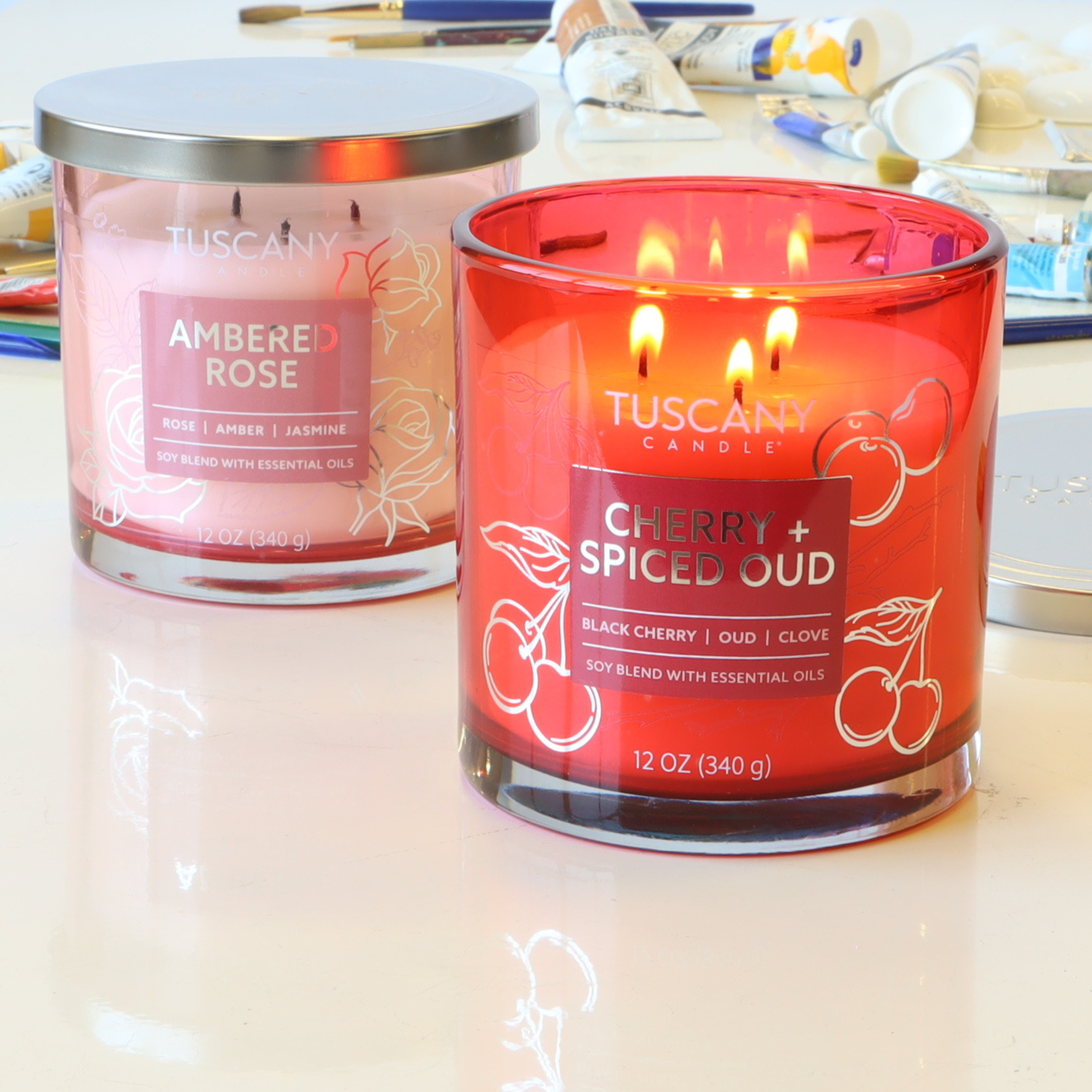 Two Tuscany Candle® EVD items: a glowing Cherry + Spiced Oud: Black Cherry Scented Jar Candle (12 oz) from the Painters' Palette Collection and an unlit "Ambered Rose," with art supplies in the background.