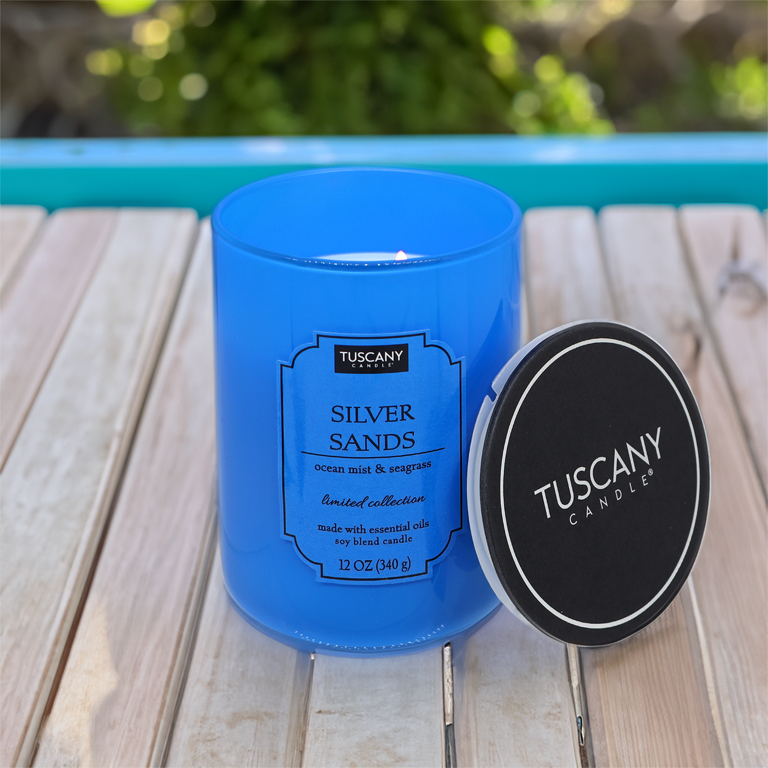 A blue Silver Sands (12 oz) candle from the Colorsplash Collection by Tuscany Candle® EVD, scented with ocean mist and seagrass fragrance, featuring a black lid on a wooden table, with clear labels.