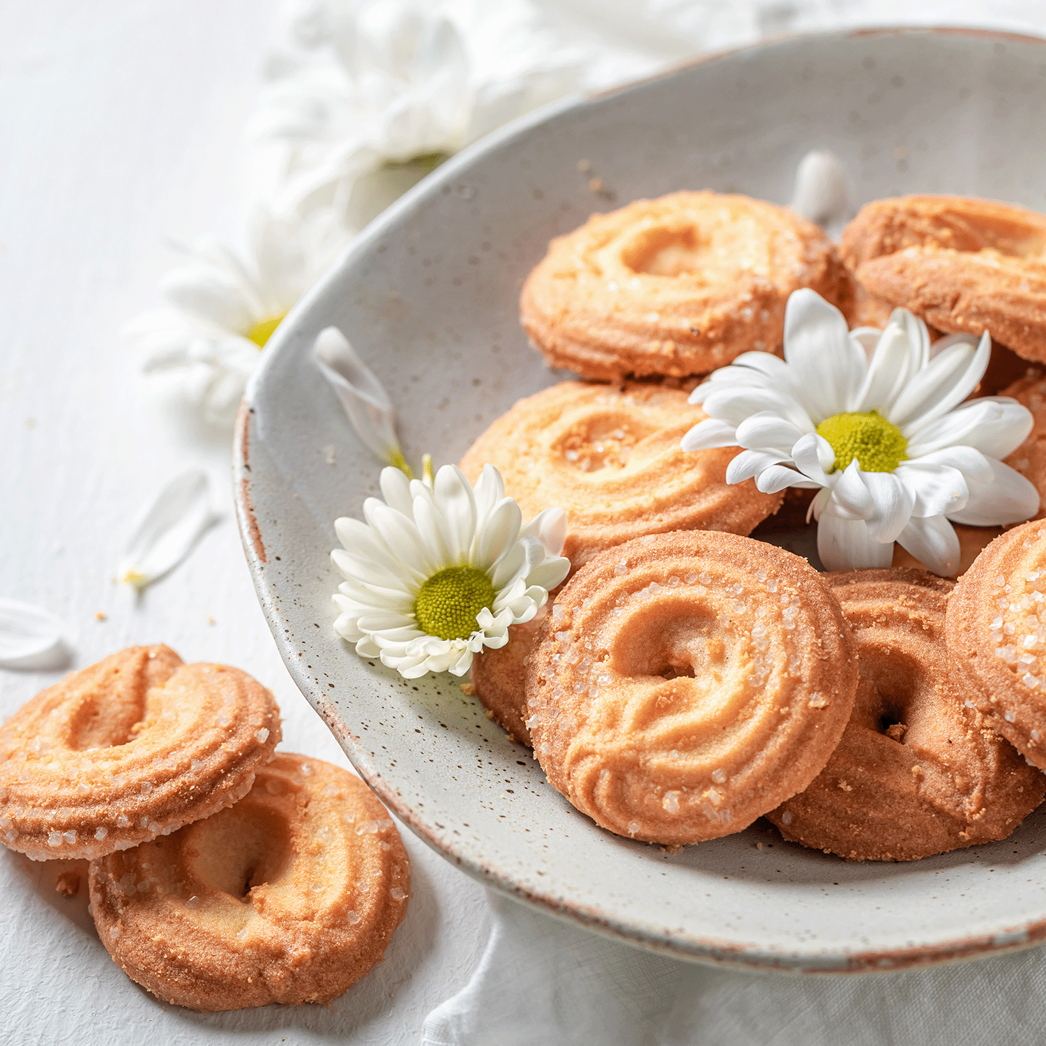 A plate of sugar-coated butter cookies is garnished with white daisies, akin to a Mother's Day celebration. Nearby, the Sweet Mom: Pistachio Shortbread Scented Candle by Tuscany Candle® SEASONAL accompanies a daisy to complete the charming scene.