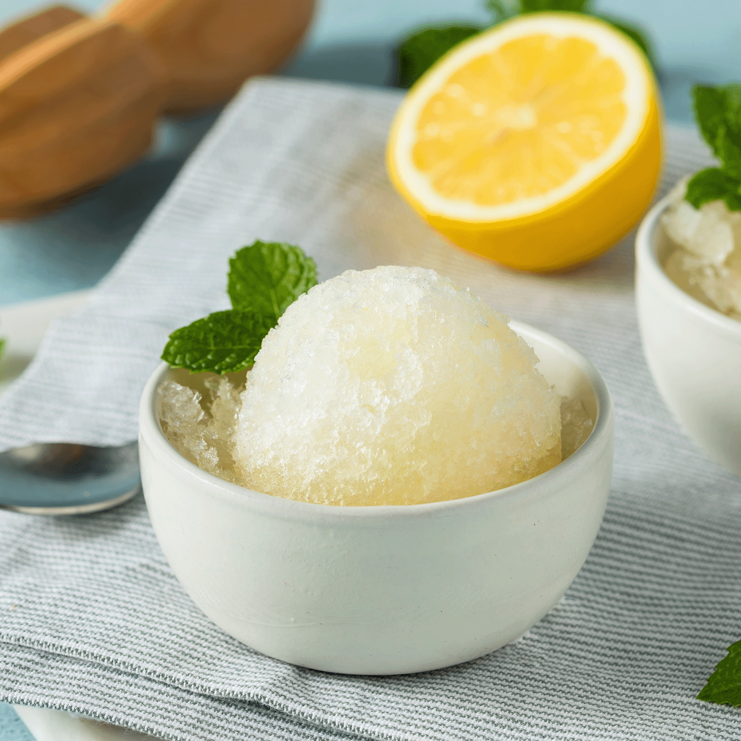 The Lemon Italian Ice: Citron & Vanilla Scented Candle (12 oz) by Tuscany Candle® SEASONAL is styled like a small white bowl of lemon granita with mint, set on a striped cloth beside a lemon half and spoon—part of the Gourmet Sorbet Collection.