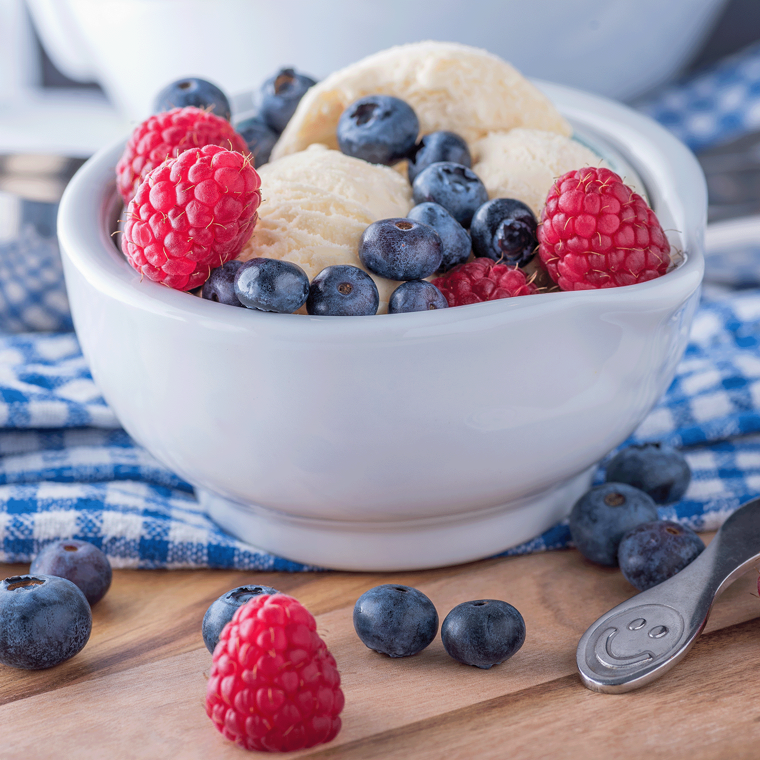 A Sour Berry Bliss: Blue Raspberry Scented Candle (12 oz) from Tuscany Candle® SEASONAL sits on a wooden surface, with its blue label and gourmet sorbet-inspired design set against a blue checkered cloth in the background.
