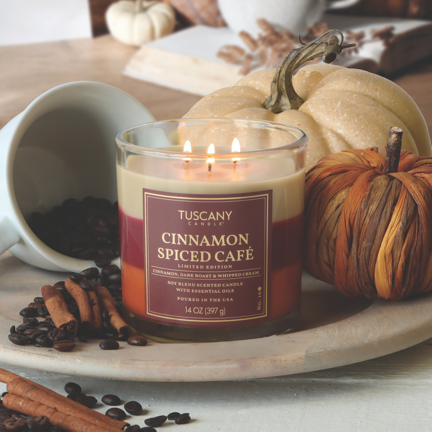 A lit Tuscany Candle® SEASONAL “Cinnamon Spiced Café: Pumpkin Spice Latte Scented Fall Candle” (14 oz) sits on a tray with pumpkins, cinnamon sticks, and scattered coffee beans.
