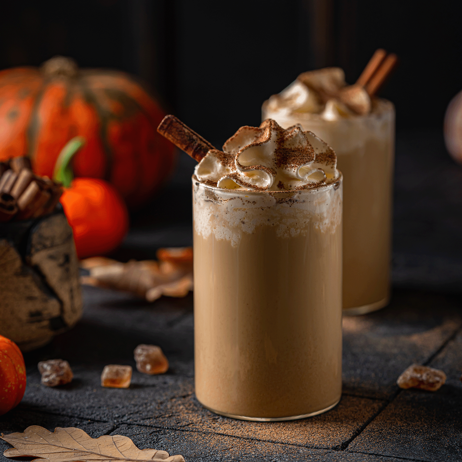 Two iced coffees with whipped cream and cinnamon sticks sit among pumpkins, leaves, ice cubes, and the Tuscany Candle® SEASONAL Cinnamon Spiced Café: Pumpkin Spice Latte Scented Fall Candle (14 oz) on a dark surface.