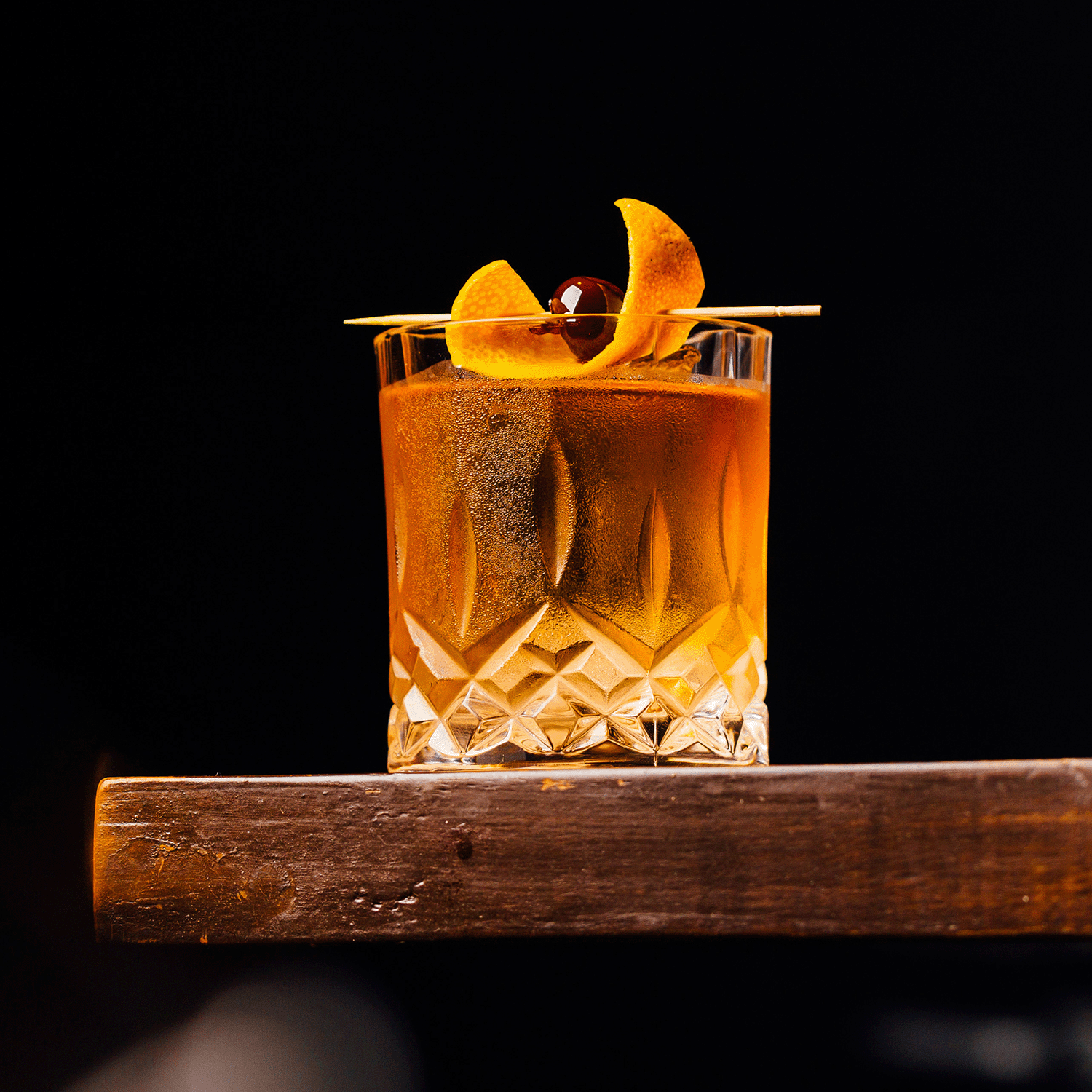 A glass of amber cocktail with ice, orange peel, and a cherry sits on wood against black, evoking the warm aroma of Tuscany Candle® SEASONAL's Bourbon Caramel Spice: Aged Bourbon & Vanilla Scented Fall Candle (14 oz).
