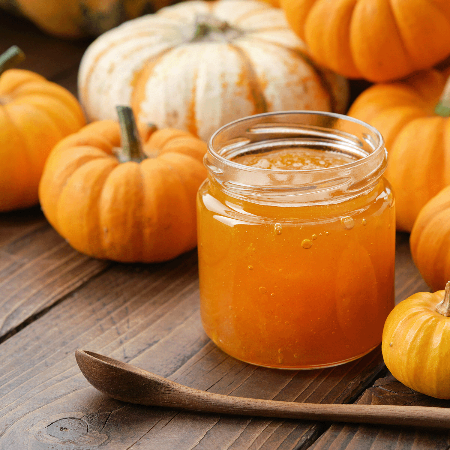 A glass jar of pumpkin jam sits on a wooden table with small pumpkins and a Tuscany Candle® SEASONAL Sugared Pumpkin: Smoked Cinnamon & Brown Sugar Scented Fall Candle (14 oz), plus a wooden spoon in the foreground.