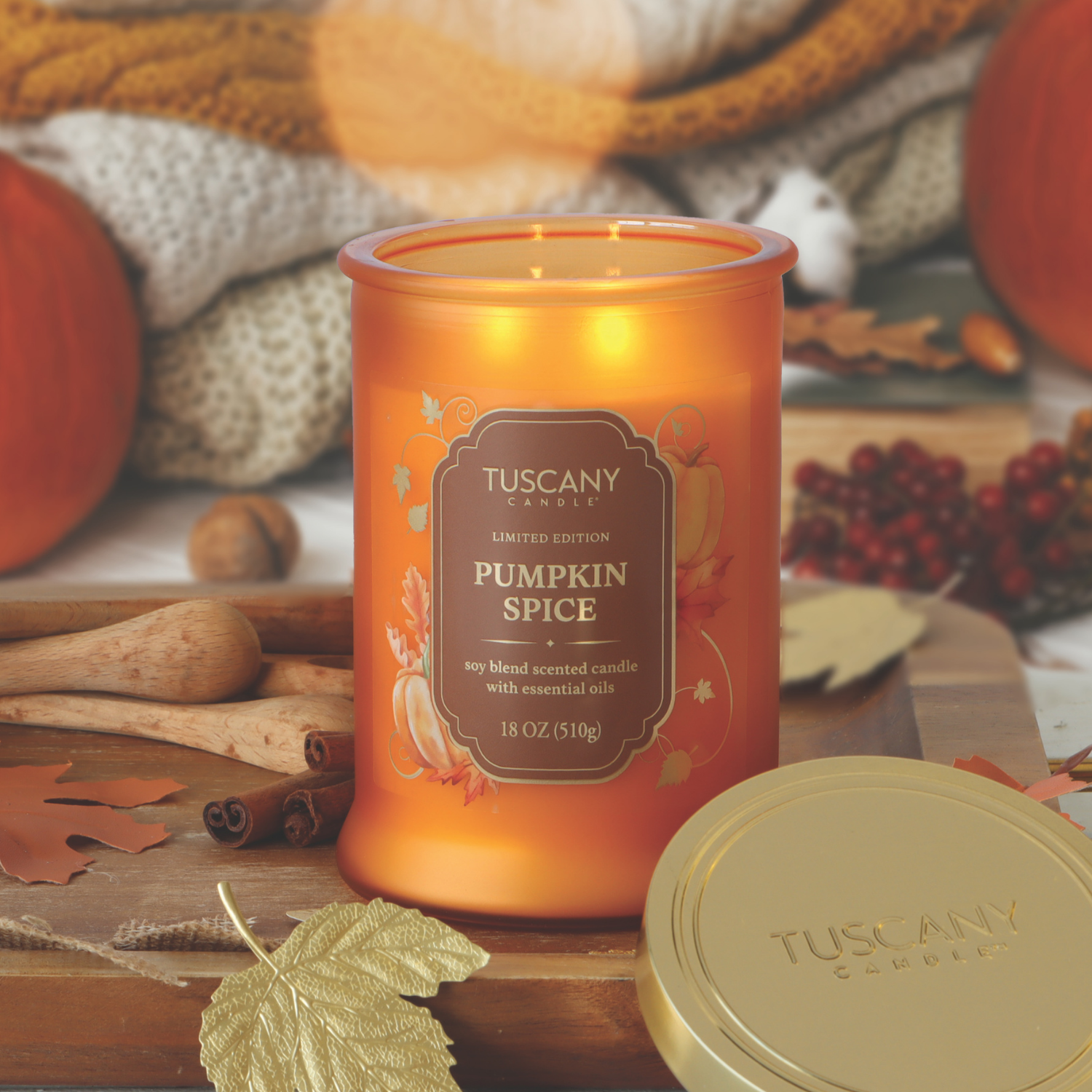 A lit Tuscany Candle® SEASONAL Pumpkin Spice: Nutmeg & Clove Scented Fall Candle (18 oz) sits on a table surrounded by fall decorations, cinnamon sticks, and cozy knit blankets.