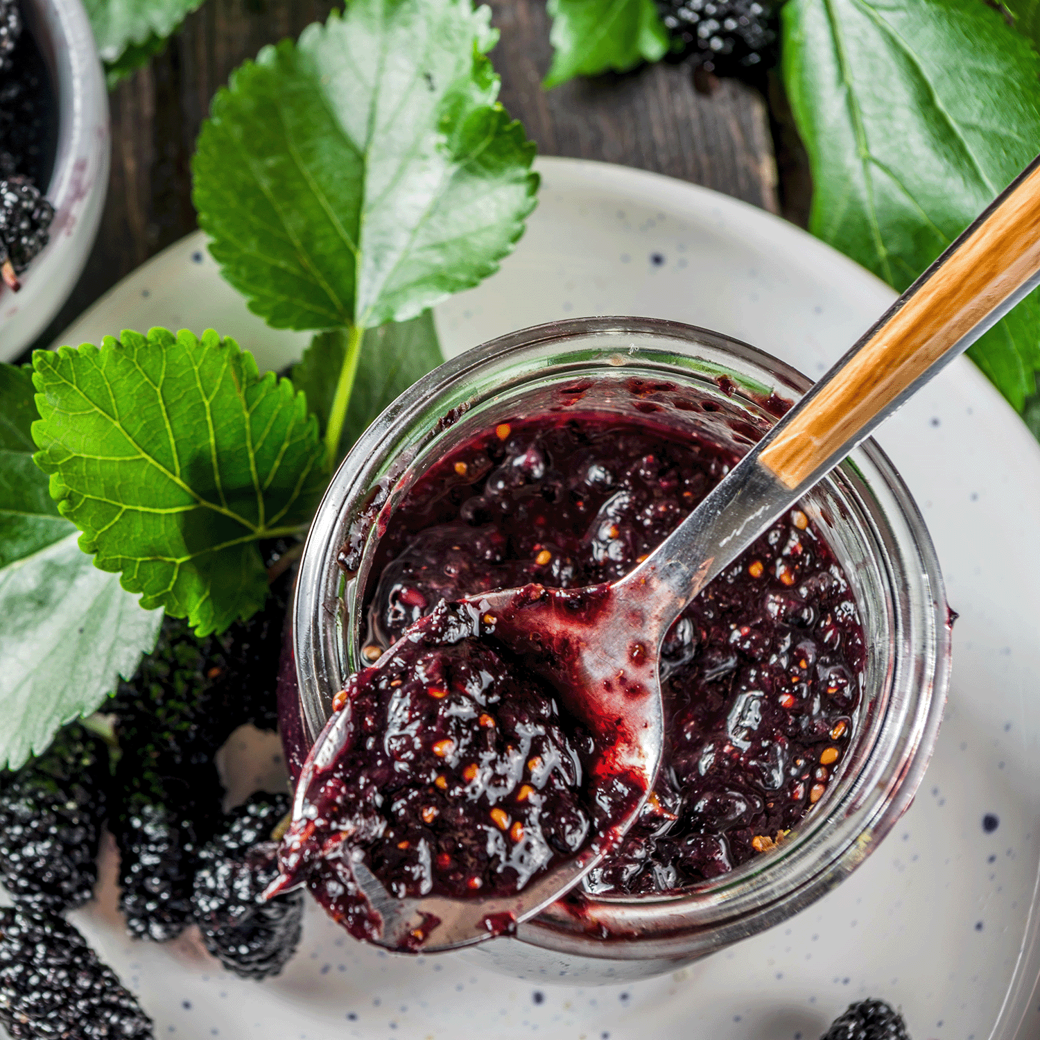 A spoon rests on a jar of dark purple jam with seeds, fresh mulberries, and green leaves—evoking the aroma of Tuscany Candle® SEASONAL's Wild Mulberry: Tart Grapefruit & Blackberry Scented Fall Candle (18 oz), made from a soy wax blend.