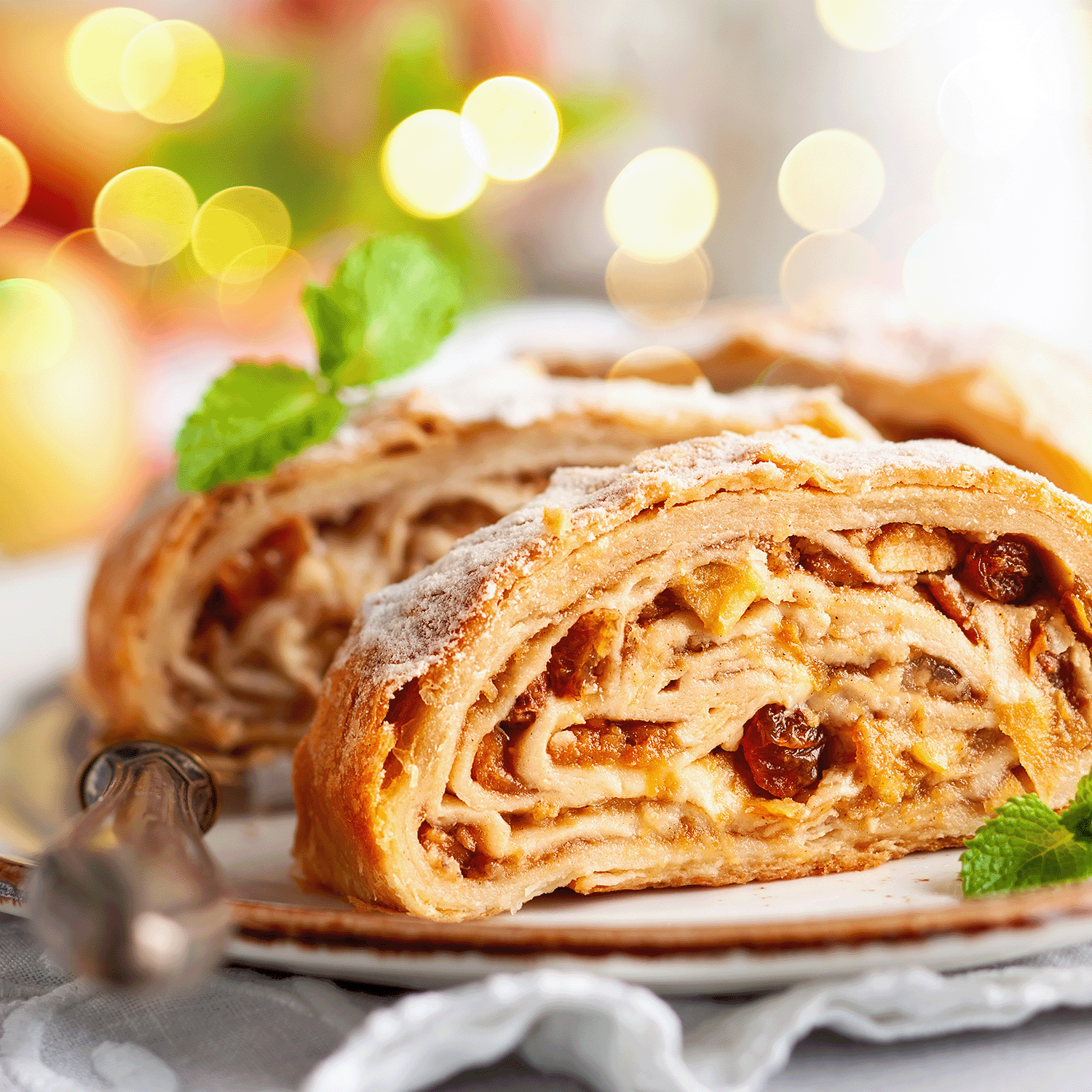 A close-up of two apple strudel slices with raisins, mint garnish, and powdered sugar conjures the cozy scent of Tuscany Candle® SEASONAL's Apple Crumble: Cinnamon & Buttercream Scented Fall Candle (12 oz).