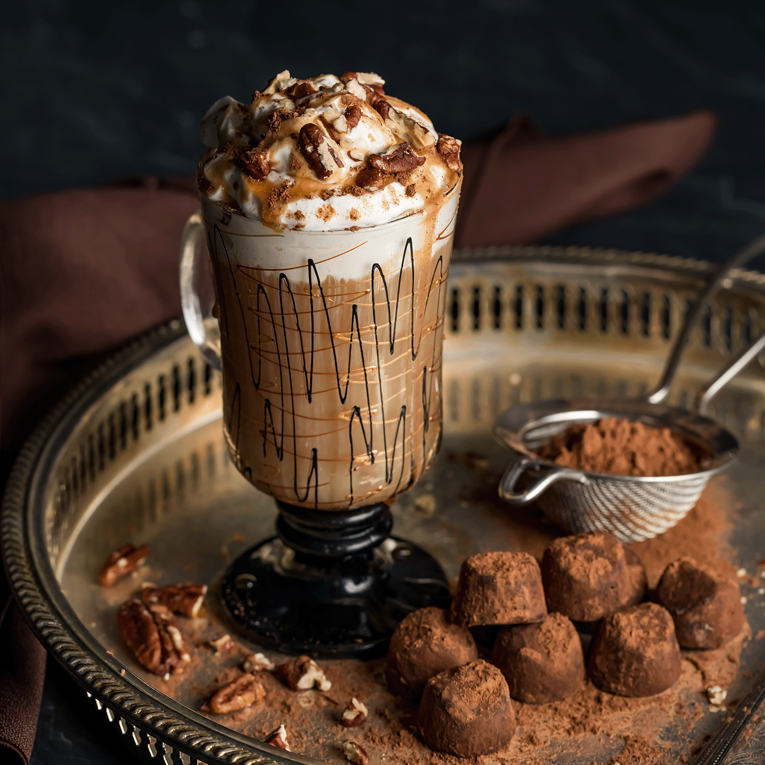 A glass of caramel nut coffee sits beside cocoa-dusted truffles and a Tuscany Candle® SEASONAL Sugarwood Molasses: Maple & Chestnut Scented Fall Candle (12 oz), filling the air with cozy autumn fragrance.