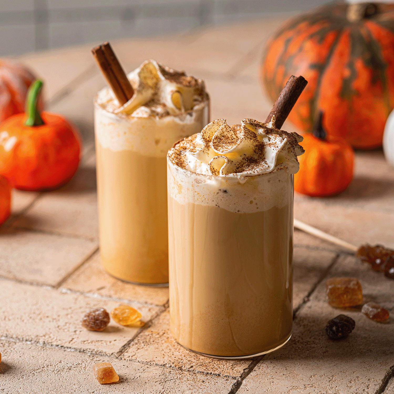 Two glasses of pumpkin spice latte with whipped cream and spice sit by pumpkins, brown sugar cubes, and a glowing Tuscany Candle® SEASONAL Pumpkin Spice Latte: Oatmilk & Dark Roast Scented Fall Candle (12 oz), creating autumn ambiance.