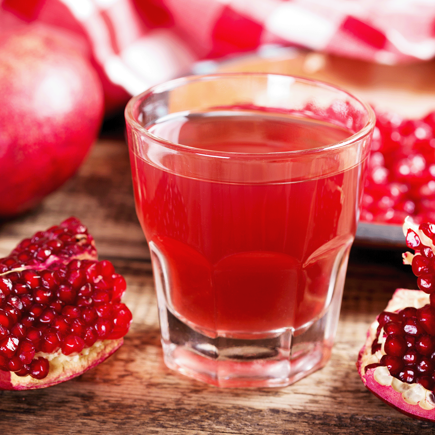 A glass of pomegranate juice rests on a wooden table beside fresh pomegranates and the Tuscany Candle® SEASONAL Apricot & Honey: Pomegranate & Patchouli Scented Fall Candle (18 oz) flickering nearby.