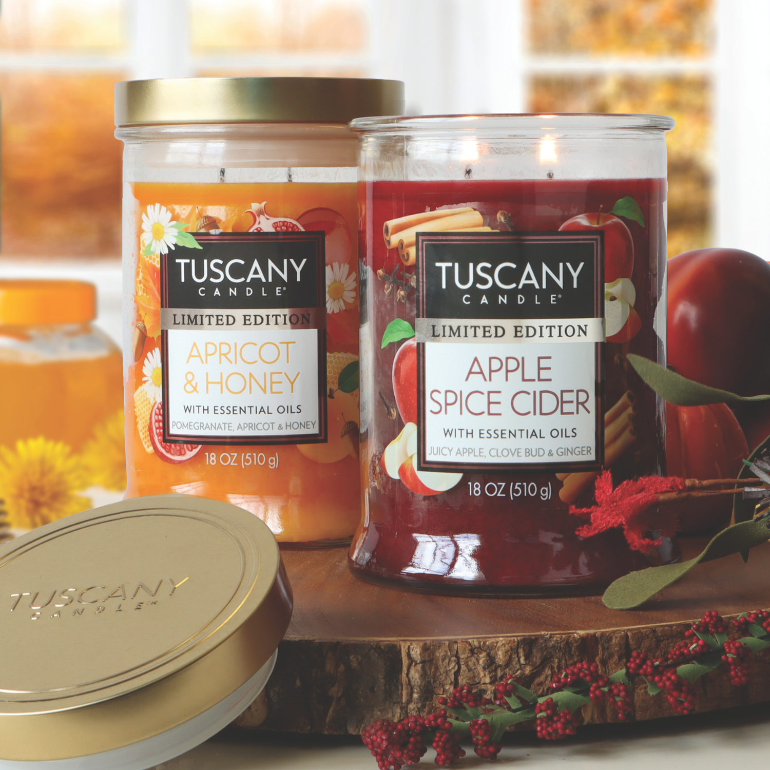 Two Tuscany Candle® SEASONAL jars—one "Apricot & Honey," the other "Apple Spice Cider: Ginger & Clove Scented Fall Candle (18 oz)"—are on a wooden surface with autumn decor. The Apple Spice Cider candle is lit.