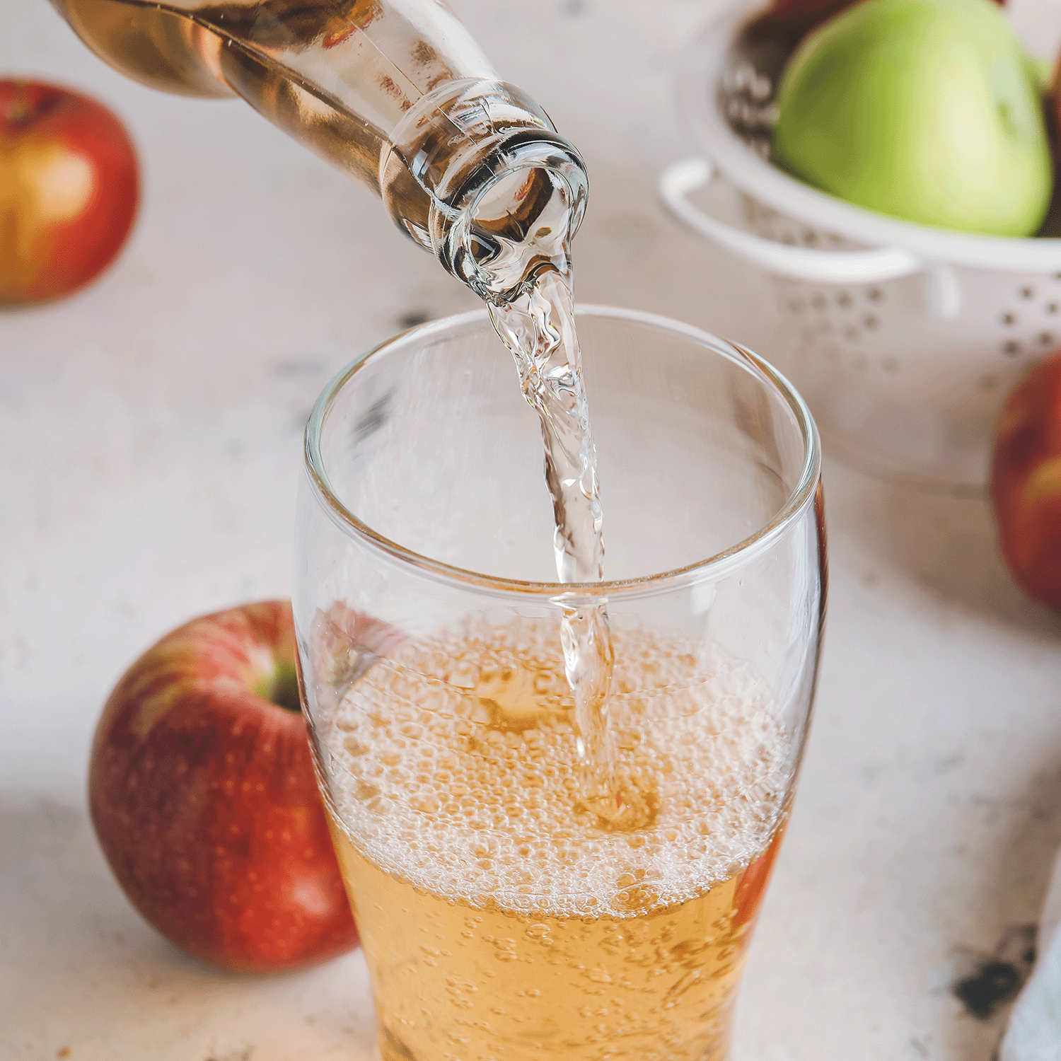A glass is filled with apple cider as red and green apples sit nearby, recalling the cozy aroma of Tuscany Candle® SEASONAL Apple Spice Cider: Ginger & Clove Scented Fall Candle (18 oz).