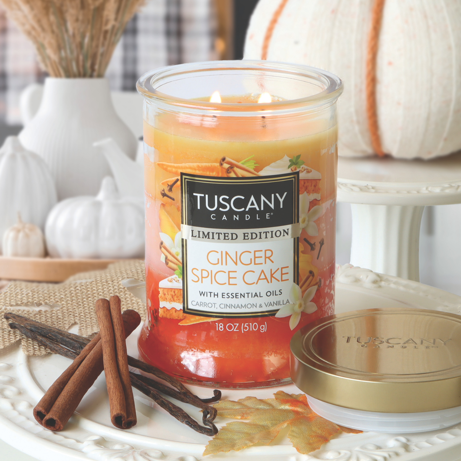 A lit Tuscany Candle® SEASONAL "Ginger Spice Cake: Carrot & Vanilla Scented Fall Candle" (18 oz) sits on a white tray with its gold lid nearby, surrounded by cinnamon sticks and white pumpkin decorations.