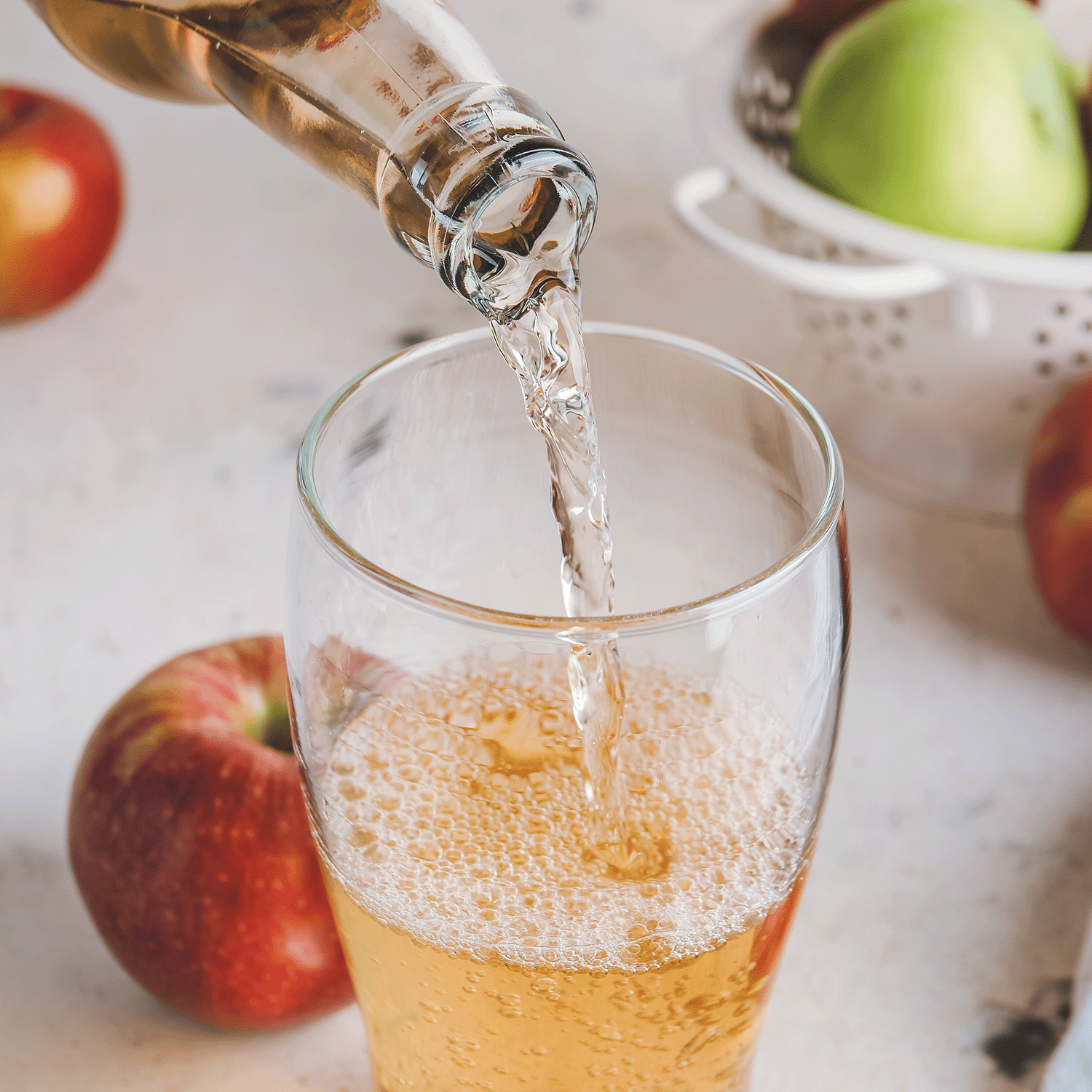 A white colander and crisp apples sit behind a glass filled with the cozy aroma of Tuscany Candle® SEASONAL Apple Spice Cider: Ginger & Clove Scented Fall Wax Melt (2.5 oz), capturing the essence of fall.