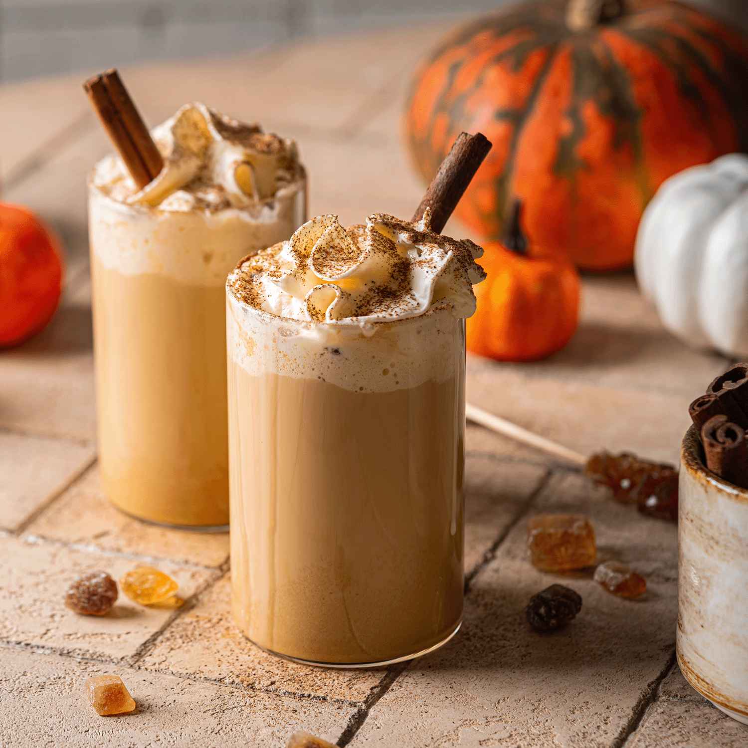 Two pumpkin spice lattes with whipped cream and cinnamon sticks sit beside a Tuscany Candle® SEASONAL Autumn Mornings: Pumpkin Spiced Latte Scented Fall Candle (14 oz), pumpkins, and sugar cubes for a cozy autumn vibe.
