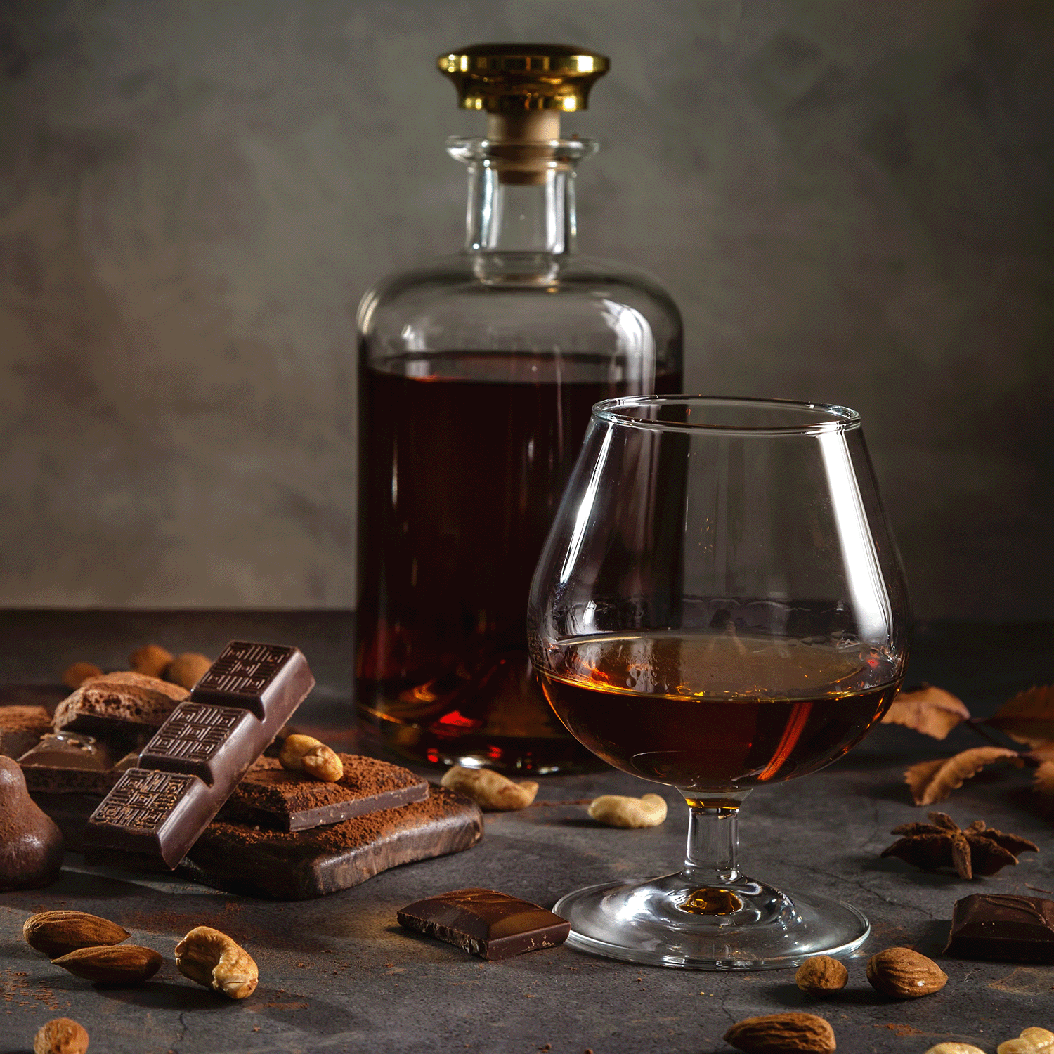A glass and decanter of brown liquor sit amid chocolate, cookies, almonds, and cashews, warmed by the glow of the Tuscany Candle® SEASONAL Bourbon Caramel Spice: Maple Sugar & Cinnamon Scented Fall Candle (12 oz) from the Bakery Tins Collection.