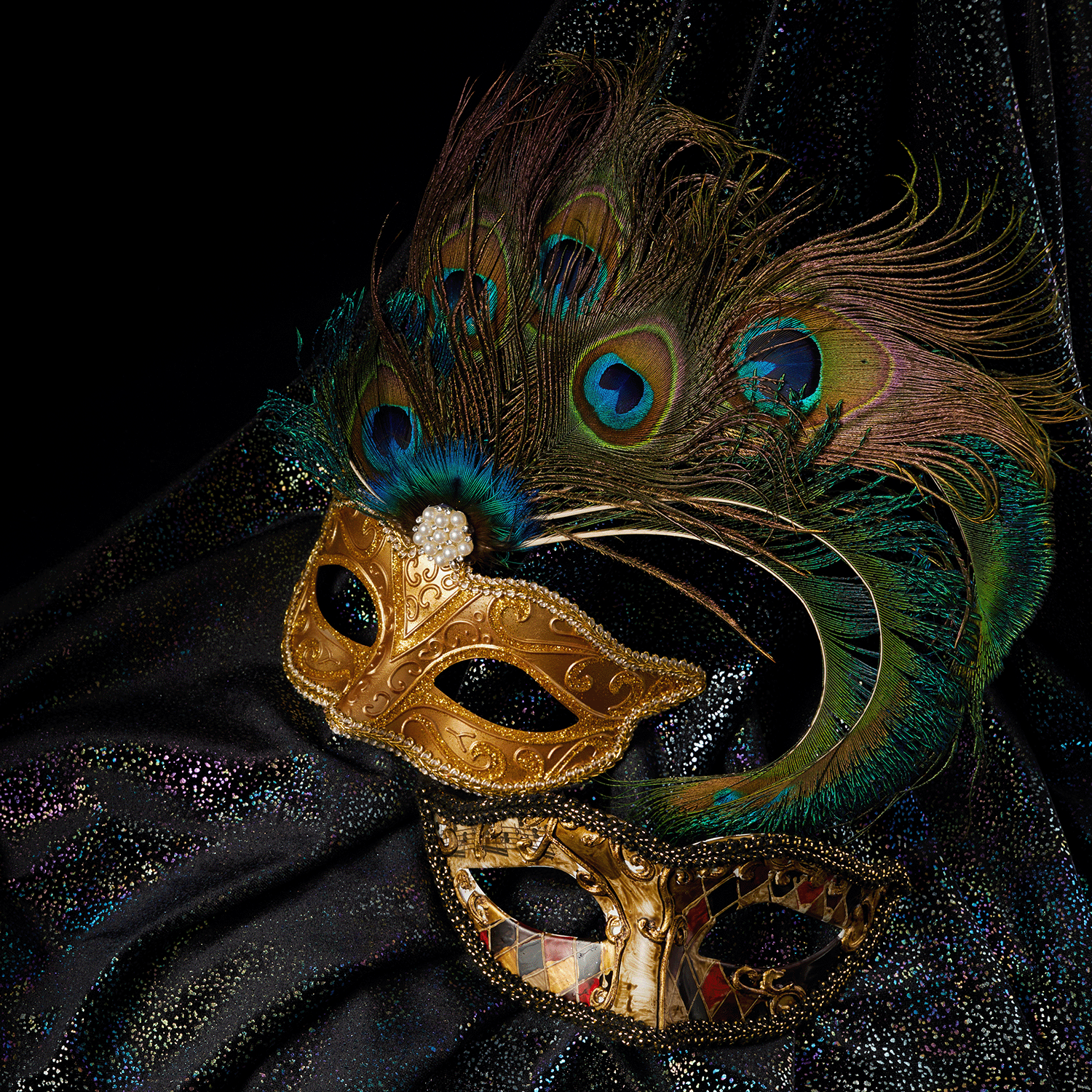 Two decorative masquerade masks—a gold one with peacock feathers and another with a diamond pattern—sit on black fabric beside Tuscany Candle® SEASONAL’s Masquerade Party Blackberry & Birch Bark Scented Halloween Candle (14 oz).