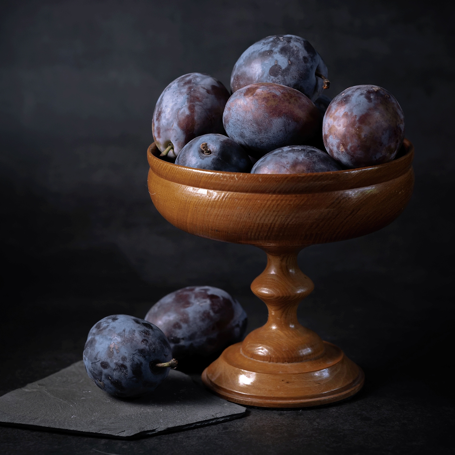A wooden pedestal bowl of dark purple plums beside two more on gray slate against a dark backdrop—perfect to display with Tuscany Candle® SEASONAL's Twilight Mist: Dark Plum & Amber Embers Halloween Candle (14 oz).