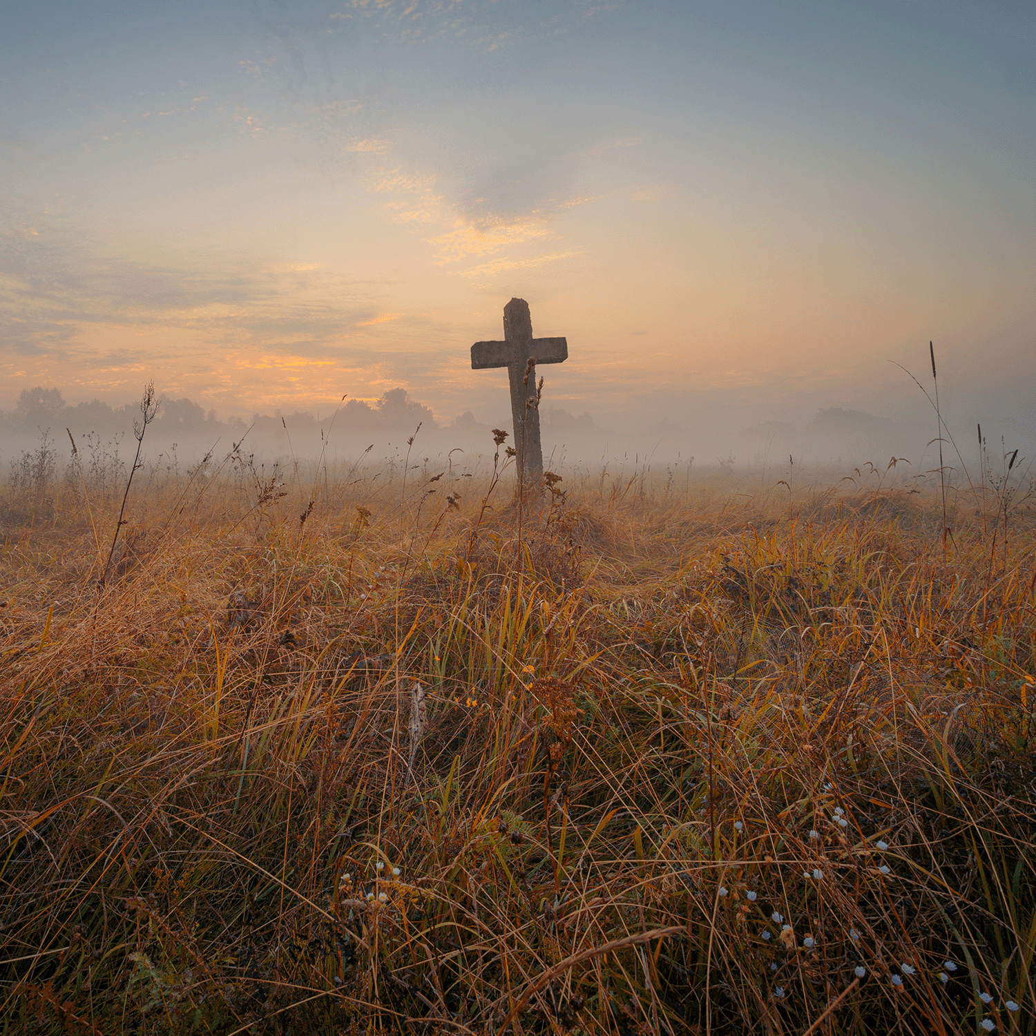 A wooden cross rises in a misty field at sunrise as the aroma of Tuscany Candle® SEASONAL’s Graveyard Fog: Crushed Leaves & Autumn Breeze Scented Halloween Candle (14 oz) drifts through autumn grass and wildflowers under a pastel sky.