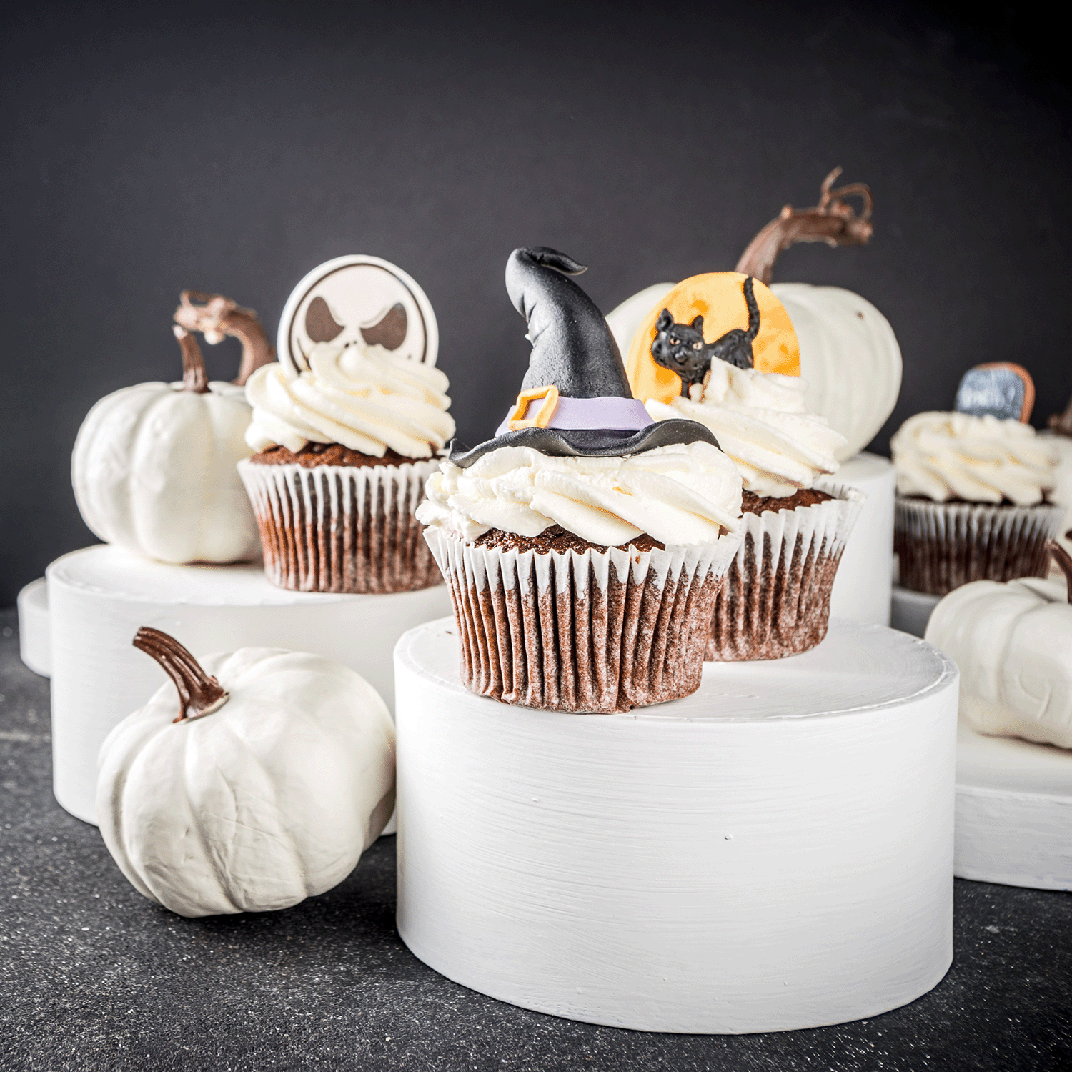 Chocolate cupcakes with white frosting and Halloween toppers are displayed on stands next to the Witching Hour: Poisoned Blueberry & Vanilla Sugar Scented Halloween Candle (Tuscany Candle® SEASONAL) and white pumpkins in the background.