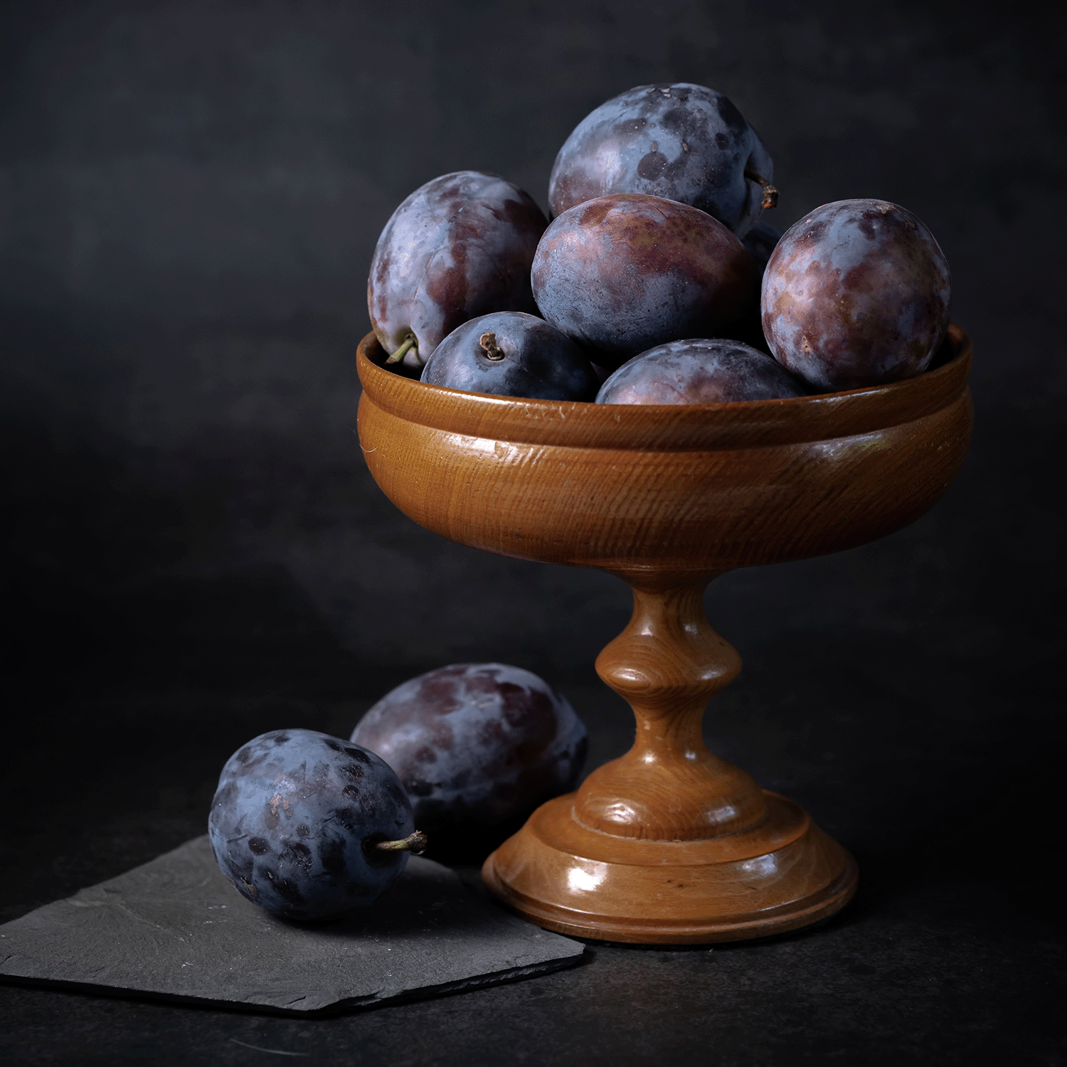 A wooden pedestal bowl filled with ripe purple plums and two plums beside it on a dark surface captures the rich hues and alluring scent of Tuscany Candle® SEASONAL Twilight Mist: Dark Plum & Amber Embers Halloween Wax Melt (2.5 oz).