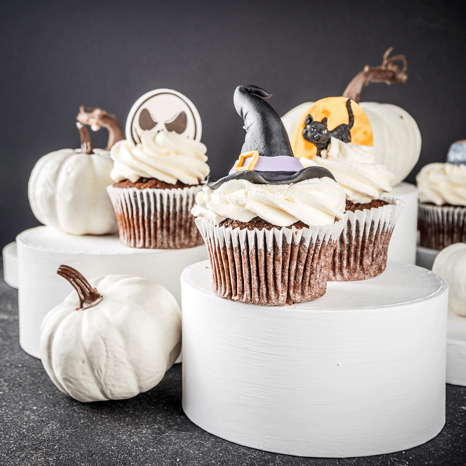 Chocolate cupcakes with white frosting are topped with Halloween decorations and displayed on white stands, surrounded by white pumpkins and Tuscany Candle® SEASONAL Witching Hour: Poisoned Blueberry & Vanilla Sugar Scented Halloween Wax Melt (2.5 oz).