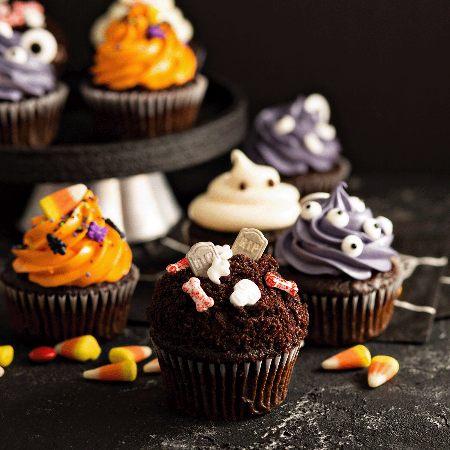 Assorted Halloween cupcakes with colorful frosting and candy are arranged beside the Spooky & Sweet: Baked Vanilla & Oatmeal Scented Halloween Candle (14 oz) from Tuscany Candle® SEASONAL, all set on a dark surface with festive candy corn.