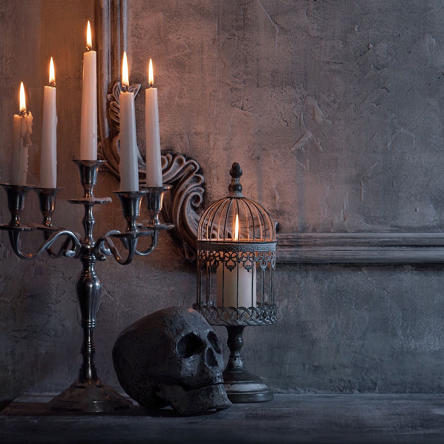 A lit candelabra, a birdcage with Tuscany Candle® SEASONAL's Mysteriously Spooky: Apple Cinnamon & Amber Scented Halloween Candle (14 oz), and a skull are displayed on a table against a textured gray wall.