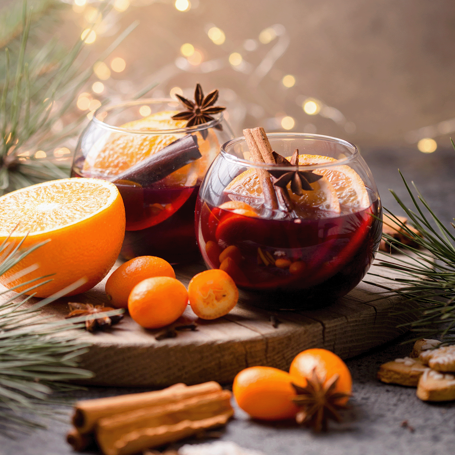 Two glasses of mulled wine with cinnamon and star anise sit among sliced orange, kumquats, pine branches, and the warm glow of Tuscany Candle® SEASONAL Hearthside Cider: Spiced Apple Cider Scented Holiday Candle (18 oz) amid festive lights.