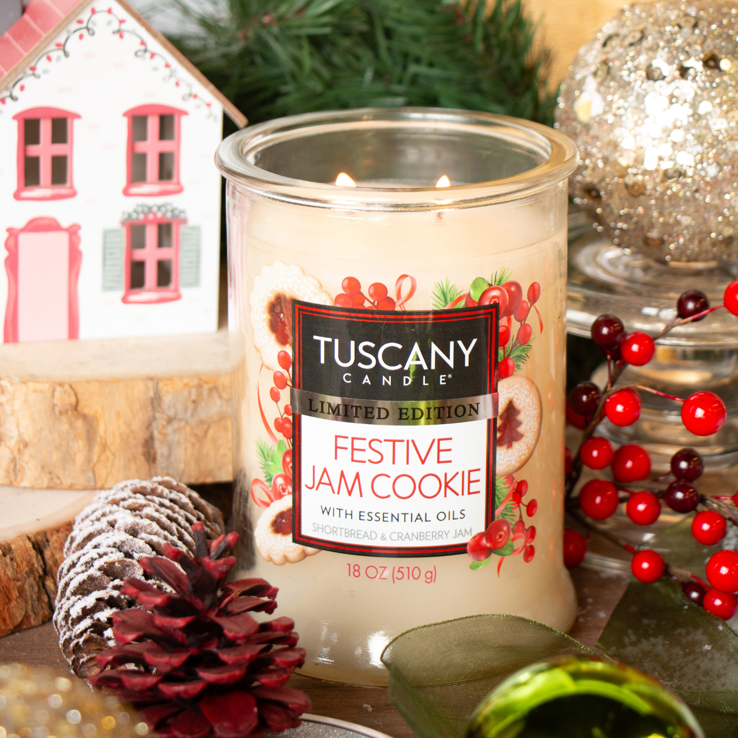 A Tuscany Candle® SEASONAL "Festive Jam Cookie: Shortbread & Cranberry Scented Holiday Candle" (18 oz) is displayed amid pinecones, red berries, greenery, and other holiday decorations.