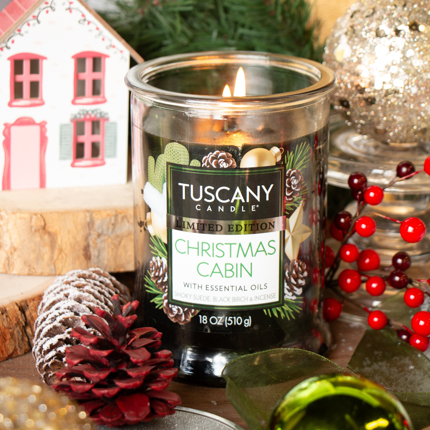 A Tuscany Candle® SEASONAL Christmas Cabin: Smoky Suede & Birch Scented Holiday Candle (18 oz) sits on a festive table with pinecones, red berries, a model house, and holiday decorations in the background.