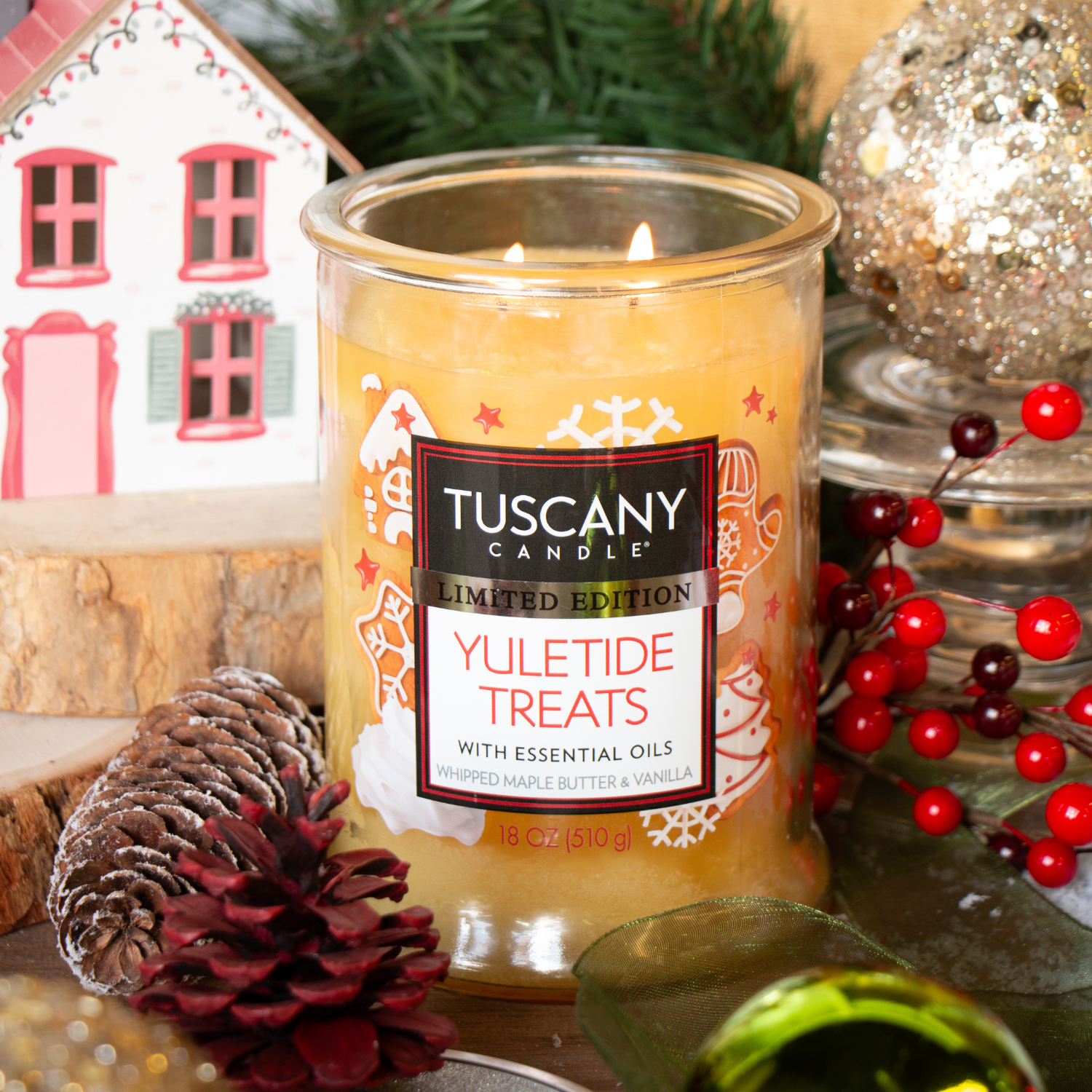 A lit Tuscany Candle® SEASONAL Yuletide Treats: Whipped Maple & Vanilla Scented Holiday Candle (18 oz) glows among pinecones, berries, greenery, and a decorative house.